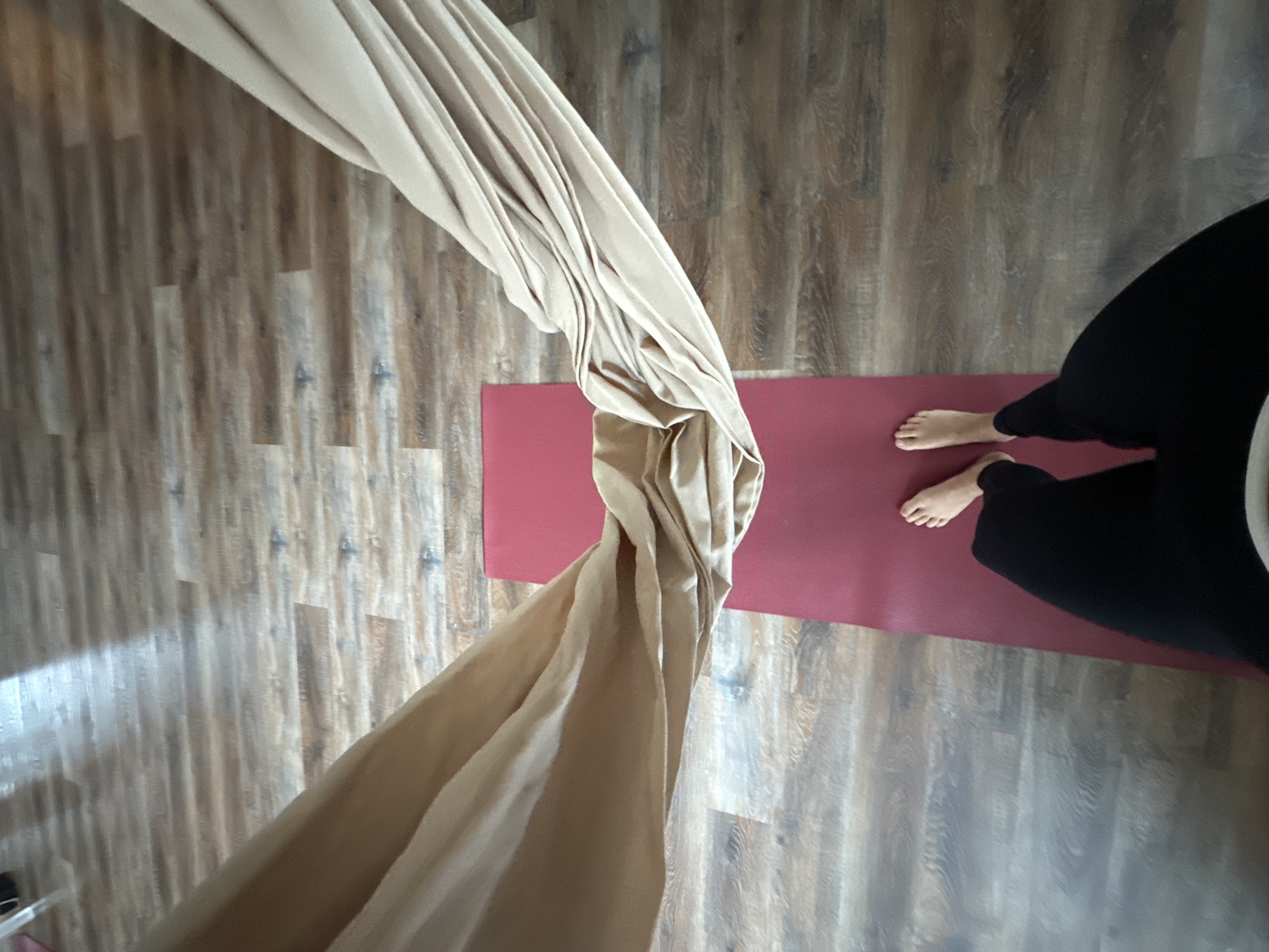 A top-down shot shows a beige aerial silk hanging over a red yoga mat on a wood-look floor. A person wearing black pants stands on the mat. The location is potentially in Krasnoyarsk, Russia, but there's no way to confirm this from the visual data alone.