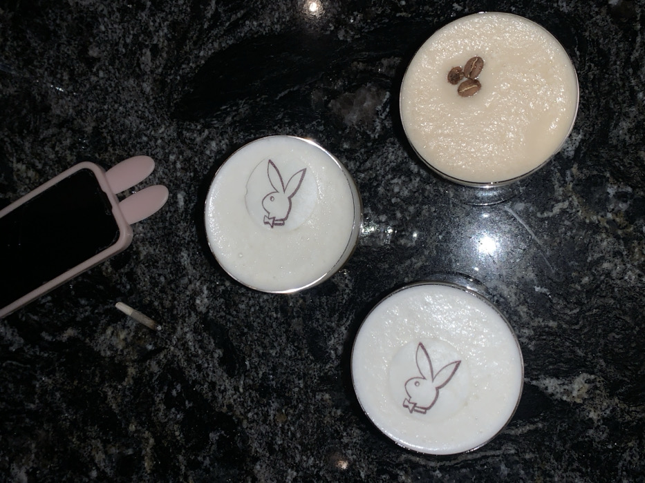 Three cocktails rest on a dark, speckled surface. Two are adorned with the Playboy bunny logo, while the third has three coffee beans on top. A mobile phone with bunny ears attached to its case lies near the drinks. A partially smoked cigarette is on the left, near the phone. The image has a dark, moody aesthetic.