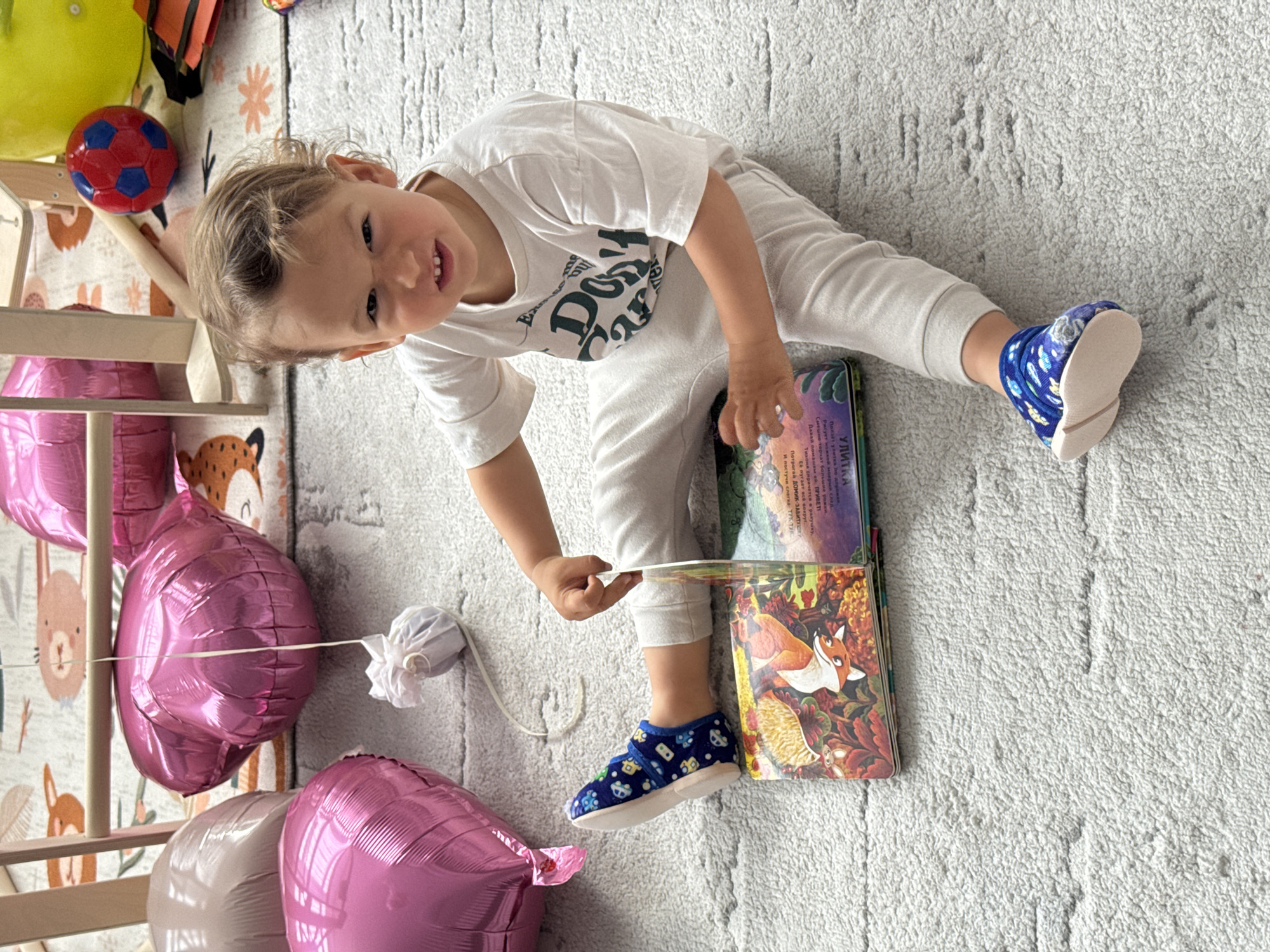 A toddler is lying on a grey carpet, propped up on their left side, and turning a page in a stack of children's books with their right hand. They are wearing a white T-shirt, grey pants, and blue patterned slippers. To the left of the child are pink balloons tied to a wooden structure. The books have colorful illustrations on their covers, including one with a fox. The setting appears to be indoors, possibly in a child's playroom. OCR text indicates a book in Russian titled "The Snail". The city of Sochi, Russia is not visible in this image.