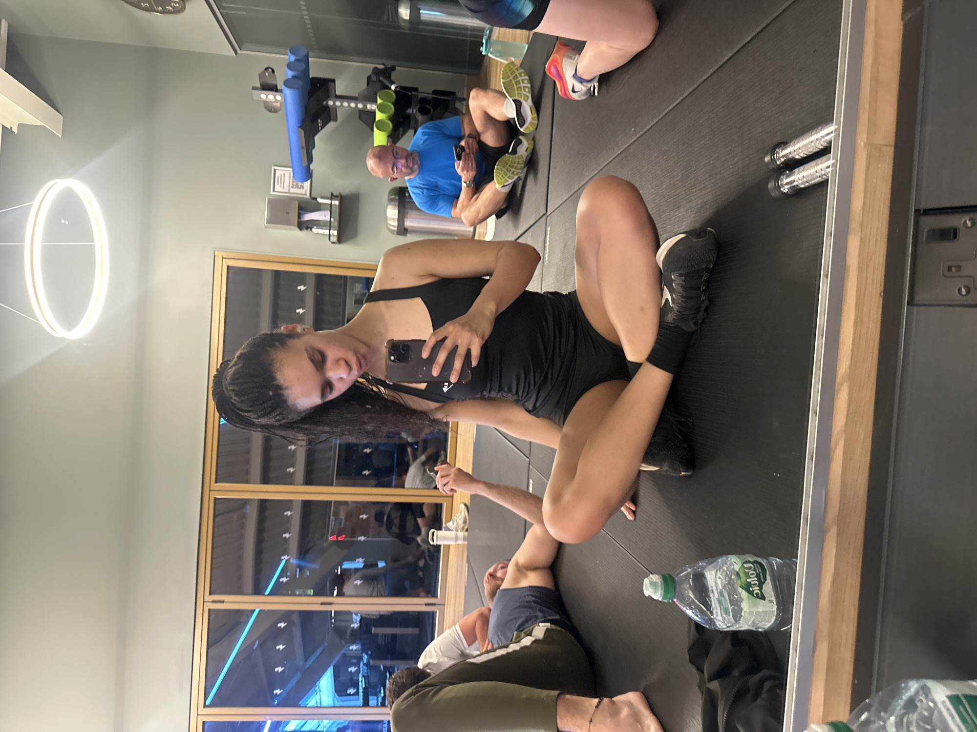 The image depicts a gym setting, likely in London, United Kingdom, given the architectural features visible through the windows. A woman in athletic wear is in the foreground, holding a smartphone and seemingly taking a selfie in a mirror. Other individuals are present, appearing to be engaged in stretching or other fitness-related activities on black mats. A man in a blue shirt and workout gear is seated on the mat, looking at his phone. Several water bottles are scattered on the floor, and fitness equipment is visible in the background. The gym has large windows, offering a view of what appears to be an urban environment.