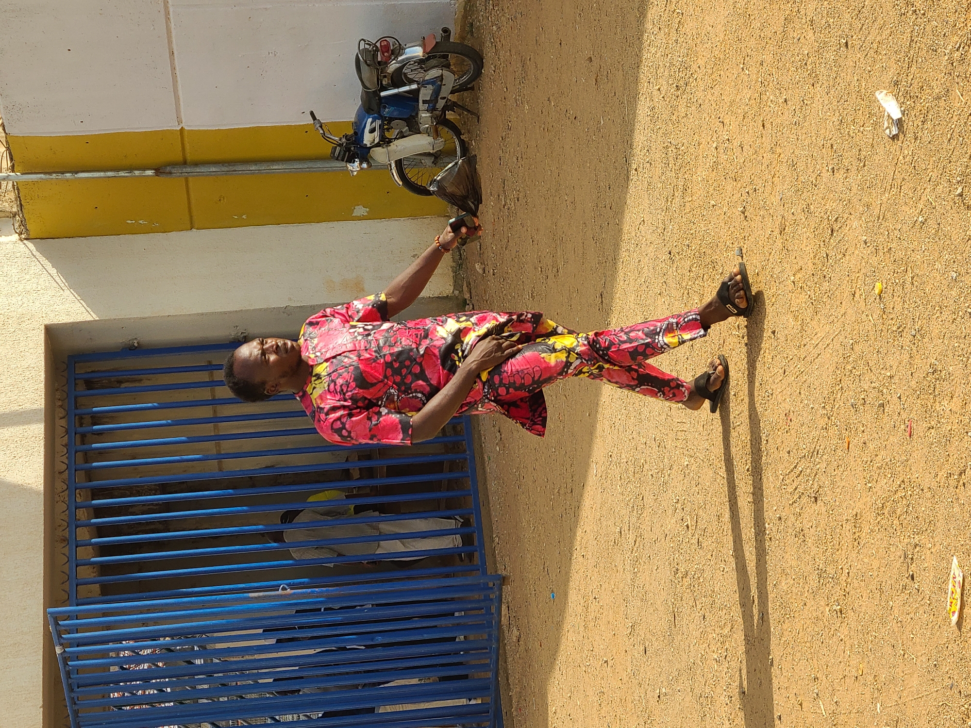 A man is standing on a dirt surface near a building in New Stadium, Nigeria. He is wearing a brightly colored red, yellow, and black patterned shirt and matching pants. He has sandals on his feet. He is holding a black plastic bag in his right hand. To the left of the man is a blue metal gate. A motorcycle is parked near the building. There is trash scattered on the ground. The building behind him is painted white and yellow.