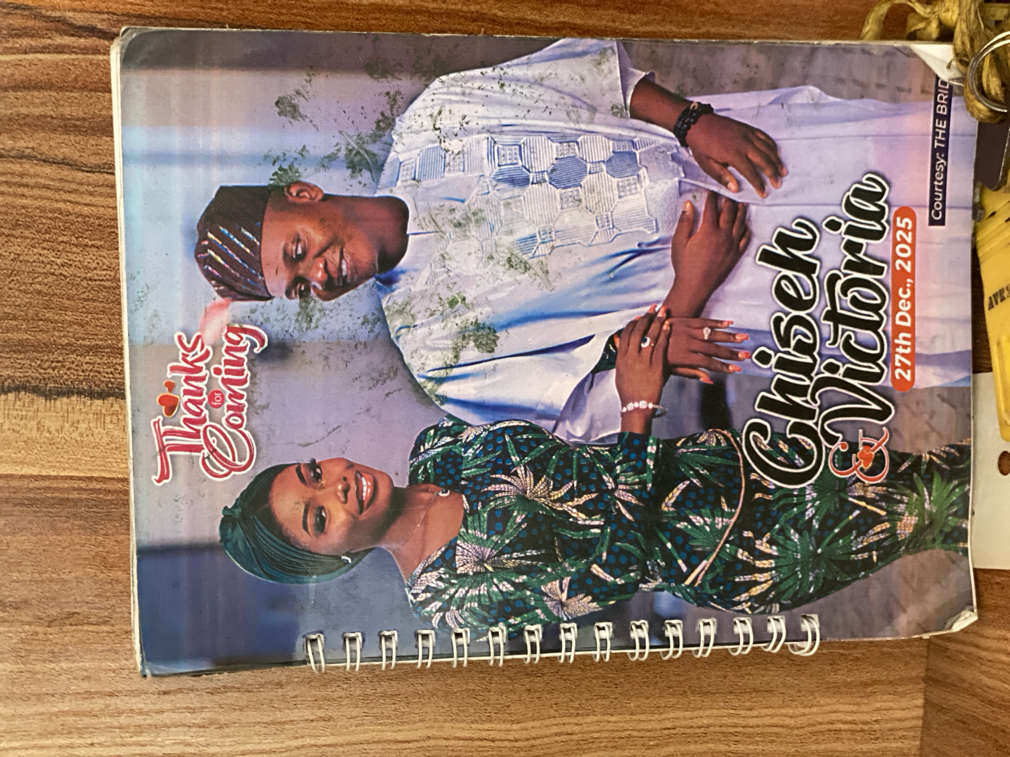 This is a photograph of the cover of a spiral-bound booklet. The cover features a posed image of a man and a woman, presumably a couple. The man is wearing a traditional light blue and white embroidered outfit and a patterned cap. The woman is wearing a vibrant green and black patterned dress and a green headscarf. Both individuals are smiling and appear happy.
The image is text-heavy. On the left side, in a cursive font, are the words "Thanks for Coming". On the right, in a bold, stylized font, are the names "Chis" and "Victoria", separated by an ampersand symbol. Below the names, in a smaller font, is the text "27th Dec., 2025". Along the right edge of the cover, a thin vertical stripe of text reads "Courtesy: THE BRID".
The background of the image is a muted, textured purple. The overall impression is a celebratory occasion, likely a wedding or a related event. The setting appears to be indoors, with the lighting suggesting daytime. The presence of the date "2025" indicates this is a pre-event invitation or acknowledgment. There are some faint green flecks, possibly from a natural element or part of the design, scattered across the cover. The photograph is taken at an angle, with a wooden surface visible beneath the booklet.