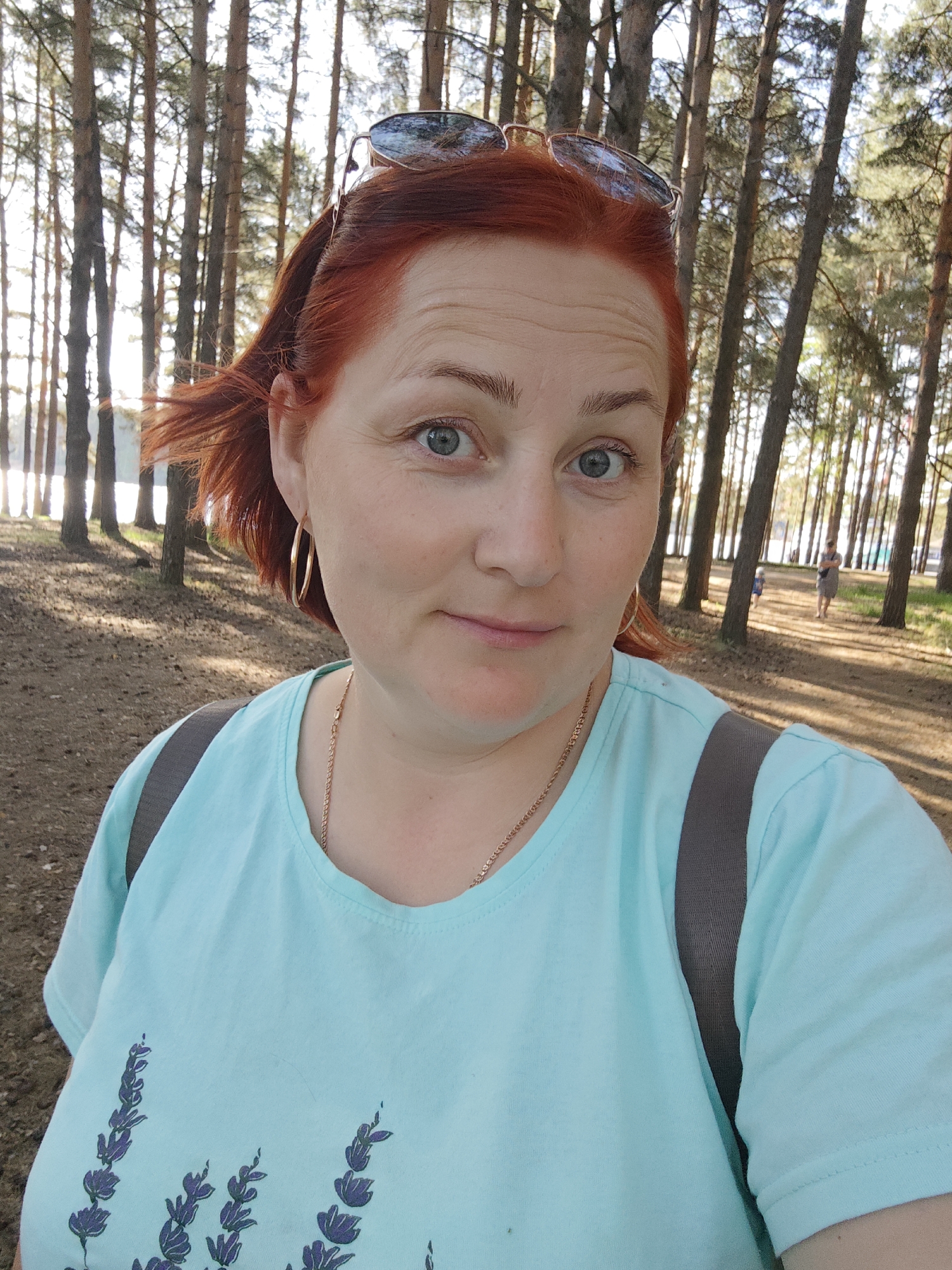 A selfie of a fair-skinned woman with red hair and blue eyes, taken outdoors in a park setting. She is wearing a light blue t-shirt with a lavender floral design, gold hoop earrings, and a gold necklace. Sunglasses are resting on top of her head. A backpack strap is visible over her shoulder. In the background, tall trees are visible, and a person is walking in the distance on a path. The photograph was taken in the city of Chaikovsky, Russia.