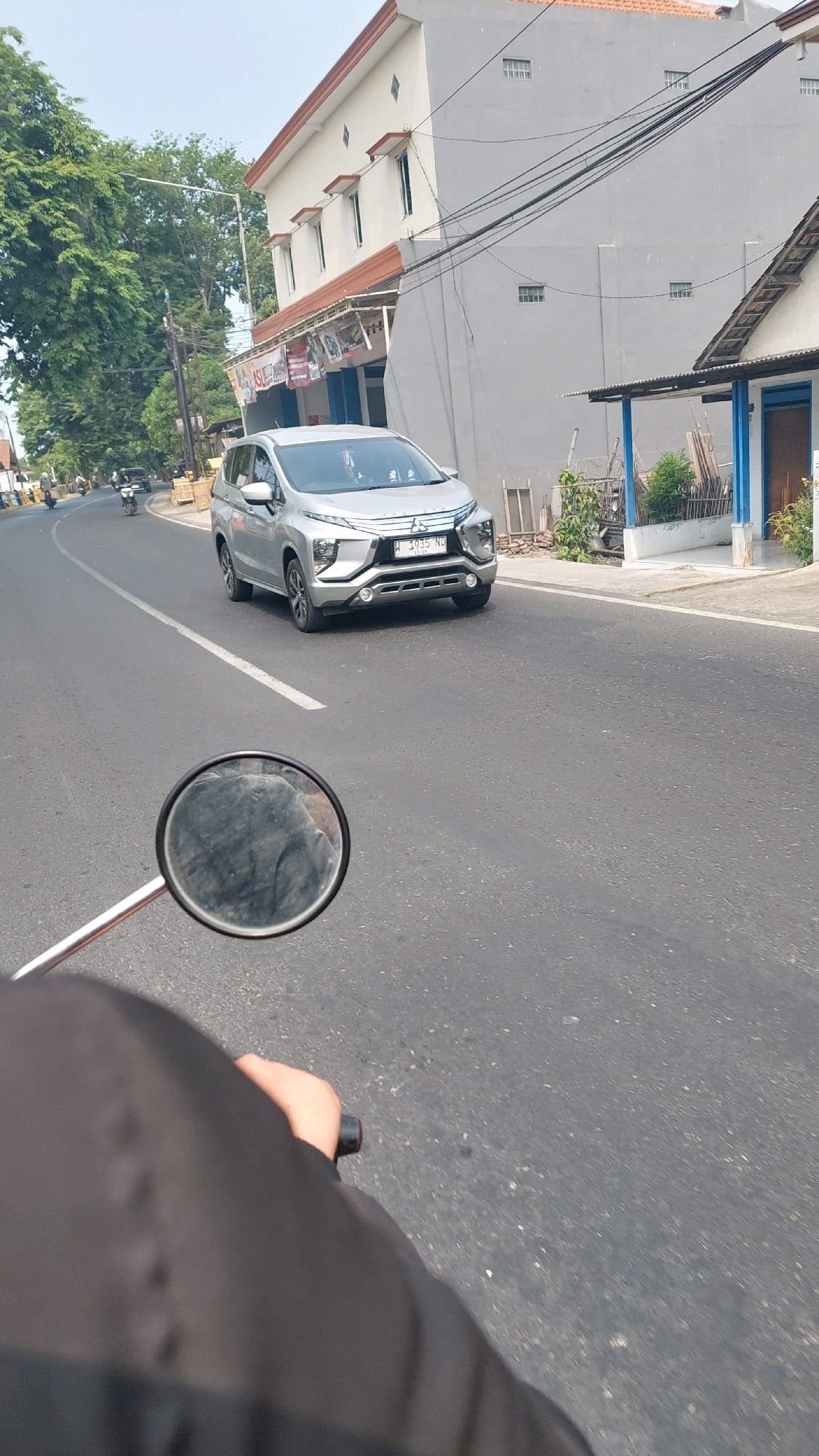 In Jimbaran Wetan, Indonesia, a silver Mitsubishi Xpander is traveling north on a two-lane asphalt road under a clear sky. The car, with license plate W 1935 ND, is positioned in the right lane. To the left of the road, a mature tree line with lush green foliage borders a residential area. On the right side of the road, a two-story building with a beige facade and red-tiled roof stands, adjacent to a single-story structure with a white facade and blue trim. The scene is captured from the perspective of a person riding a motorcycle, with the motorcycle's handlebar and a partial view of the rider's black jacket and hand visible in the foreground. The motorcycle's side mirror reflects the rider and a portion of the road. Several other motorcycles are visible in the distance on the left side of the road, heading in the same direction as the Xpander.