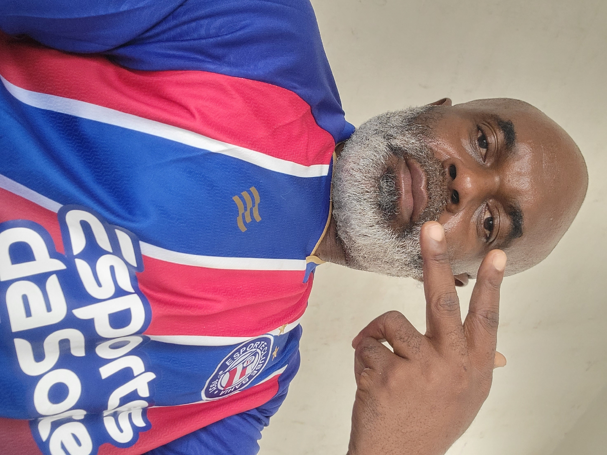 A dark-skinned man with a gray beard and bald head is pictured wearing a blue, red, and white soccer jersey with the word "ESPORTS" on it and a crest on the right side. He is making a peace sign with his right hand and looking at the camera. The background appears to be a plain, light-colored wall. No indicators of the city of Maiburiji, Nigeria can be determined from the image.