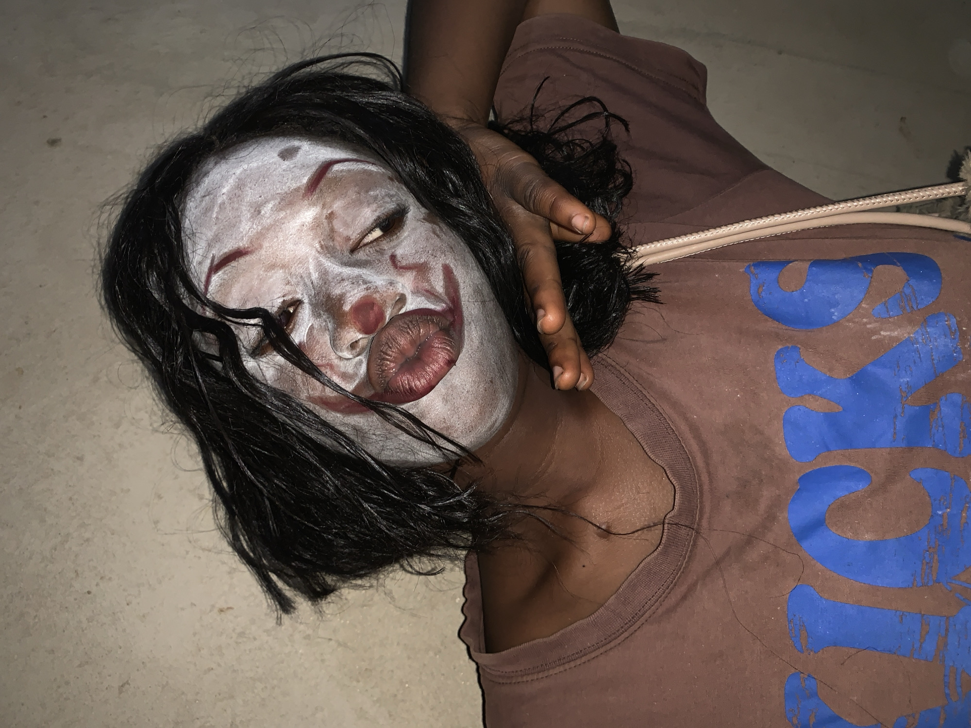 The image depicts a person lying on a light-colored surface, possibly a floor. The individual is wearing clown-like makeup, with a white base, red accents around the eyes and mouth, and dark hair. They are wearing a brown t-shirt with a large blue graphic design. One hand is near their face, touching the hair. The scene appears to be indoors, lit by an unseen light source. It is not possible to determine the exact location within Agban, Nigeria, from the image alone. The person appears to be either unconscious or posing.