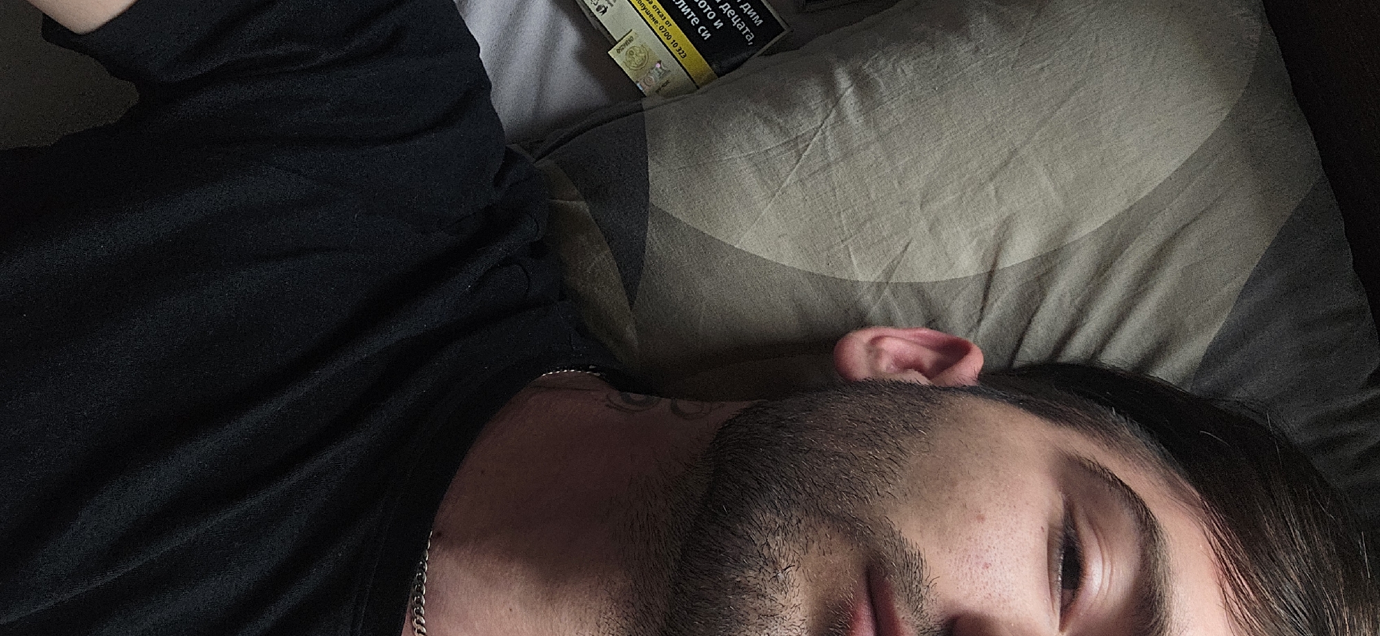 A person, likely male due to visible facial hair, is depicted lying down, possibly resting or sleeping, with their eyes partially closed. They are wearing a black long-sleeved garment and a silver chain is visible around their neck. The person's head rests on a light-colored pillow with a subtle dark grey curved pattern. Their ear and dark hair are visible against the pillow.
In the upper part of the frame, near the person's head, a yellow and black pack of cigarettes is visible on the pillow. The image is taken from a slightly overhead perspective in an indoor setting, possibly a bedroom. The lighting appears subdued.
Visible Cyrillic text on the cigarette pack includes fragments of a health warning such as "дим" (smoke), "децата," (children,), "ото и" (and), "лите си" (their), "а отказ от" (and refusal from), and a helpline number: "Попушене: 0700 10 323" (Smoking: 0700 10 323). This text is consistent with product labeling in Bulgaria.