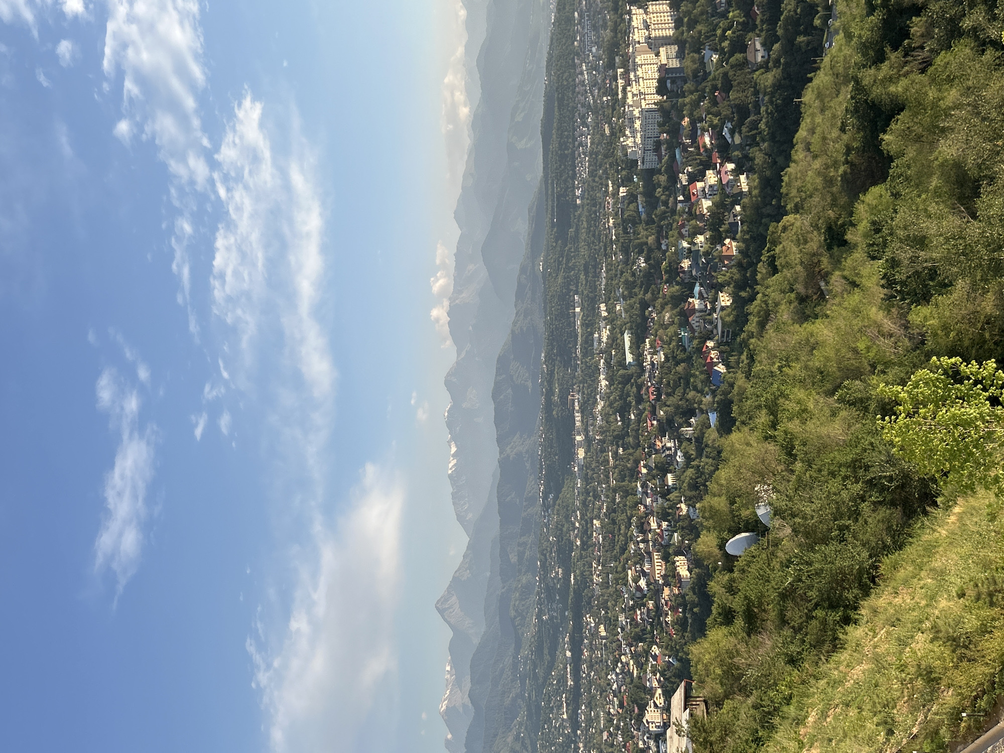 The image depicts a panoramic view of Almaty, Kazakhstan, with a city sprawled along the foothills of a majestic mountain range. The perspective is from a high vantage point, looking down and across the urban landscape, which is interspersed with lush green trees. In the foreground, the slope is covered with dense green vegetation, including bushes and grass, suggesting a natural, undeveloped area contrasting with the city. Two satellite dishes are visible amidst the trees in the mid-ground, indicating communication infrastructure.
The city itself is a mix of architectural styles, with modern, multi-story buildings and smaller residential houses, many featuring colorful roofs. The dense greenery weaves through the urban fabric, creating a harmonious blend of nature and city life. Beyond the city, the imposing mountains rise, their peaks shrouded in a light haze, hinting at a vast, natural expanse. Some of the higher mountain peaks show hints of snow, suggesting significant elevation.
The sky above is a clear blue, punctuated by scattered white clouds, indicating fair weather. The lighting suggests it is daytime, likely mid-morning or afternoon, with the sun casting a gentle illumination over the scene. There is no discernible activity of people, nor any specific event taking place. The overall mood is one of tranquility and awe, capturing the striking natural beauty that surrounds and integrates with the urban environment of Almaty. No visible text is present in the image.