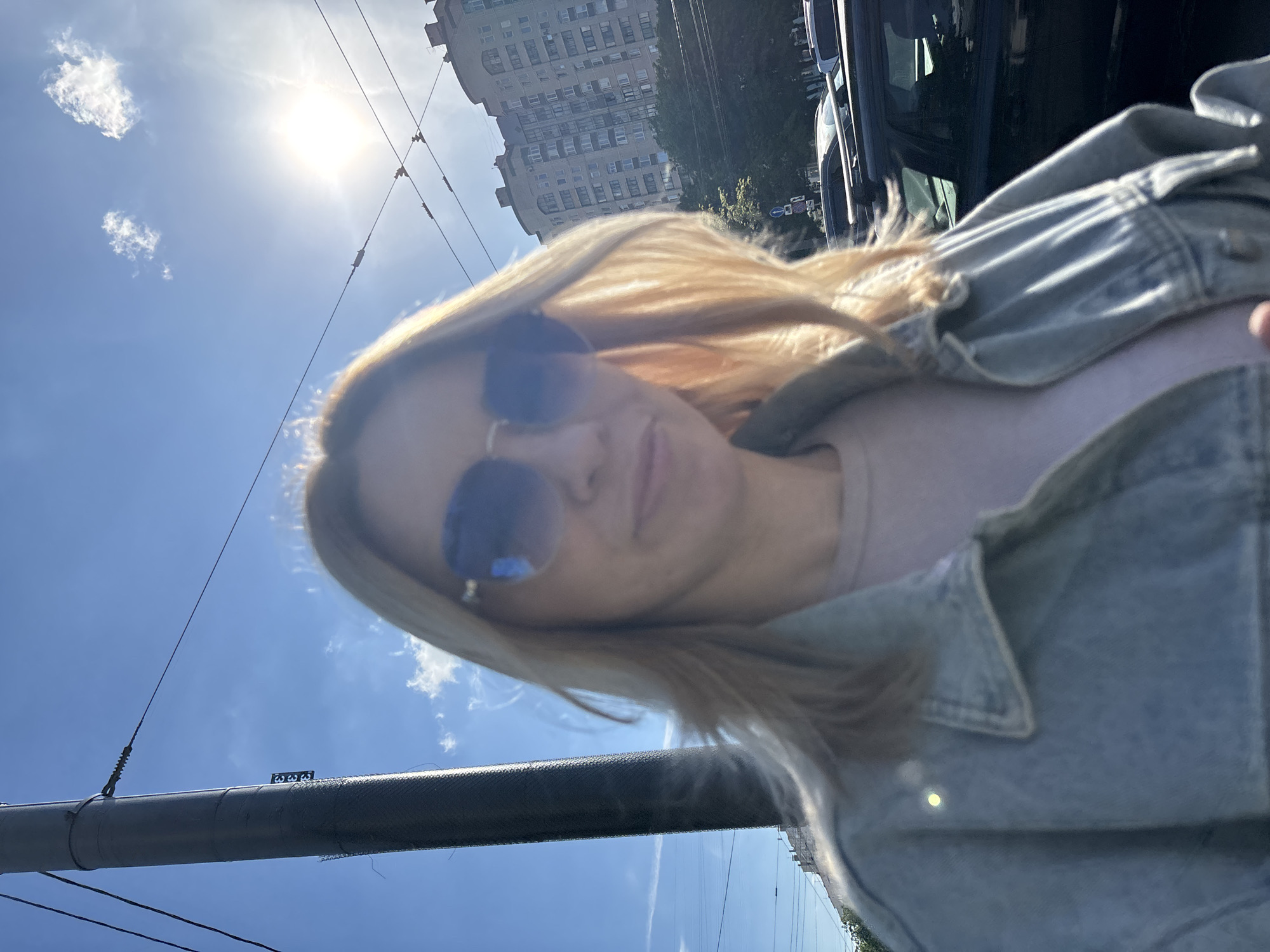 The brightly lit image is a casual outdoor selfie featuring a woman on a sunny day in an urban or suburban setting. The photograph is significantly rotated clockwise, placing the sky on the left and the ground elements on the right.
The primary subject is a woman, partially visible in the center-right of the frame. She has blonde or light-colored hair and is wearing a light blue denim jacket over a lighter-colored top, possibly pink or light purple. Her face is obscured by large, dark sunglasses with a blue or purple tint, reflecting some light. Her expression is not discernible due to the sunglasses and the angle.
The background reveals a clear, vibrant blue sky dotted with a few small, white clouds. The sun is intensely bright and positioned in the upper-left area of the rotated image, creating a strong glare. Numerous dark power lines or trolley wires crisscross the sky, suggesting the presence of urban infrastructure or public transport.
On the right side of the rotated image, a tall, multi-story building with numerous windows is visible, indicating a city environment. Dark-colored vehicles are partially seen parked or driving near the building, with some green foliage from trees or bushes also present. A large, dark utility pole or beam extends horizontally across the lower part of the image, close to the woman.
The overall impression is that of a bright, sunny day. No discernible text is visible within the image.