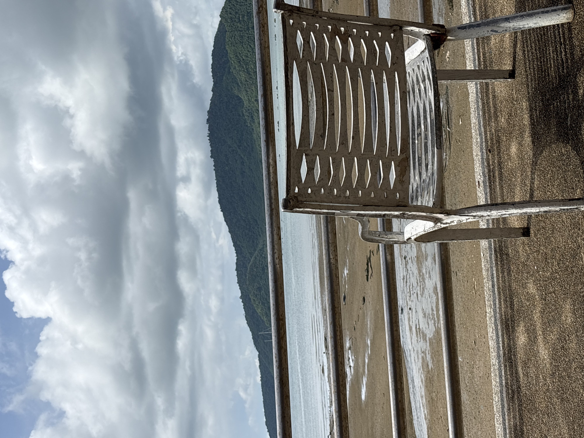 An empty, weathered white metal chair sits facing a tranquil bay with a lush green mountain in the background. The scene is located on a paved promenade in Rawai, Thailand, overlooking a beach with the tide out. The sky is filled with dramatic, cloudy formations. A metal railing separates the promenade from the beach.