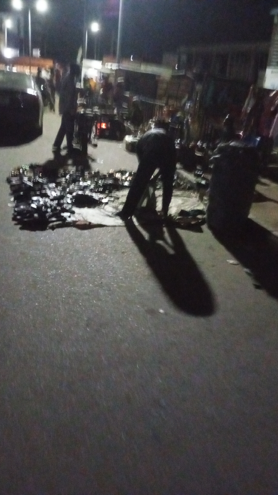 The scene depicts an outdoor marketplace at night in Lokoja, Nigeria. A large quantity of goods, possibly footwear, is displayed on the ground. Several individuals are present, some examining the items for sale, others standing nearby. A car is partially visible on the left side of the frame. Streetlights illuminate the area. The ground appears to be paved, likely a road or pedestrian area. Shadows are cast by the light sources. The environment appears urban.