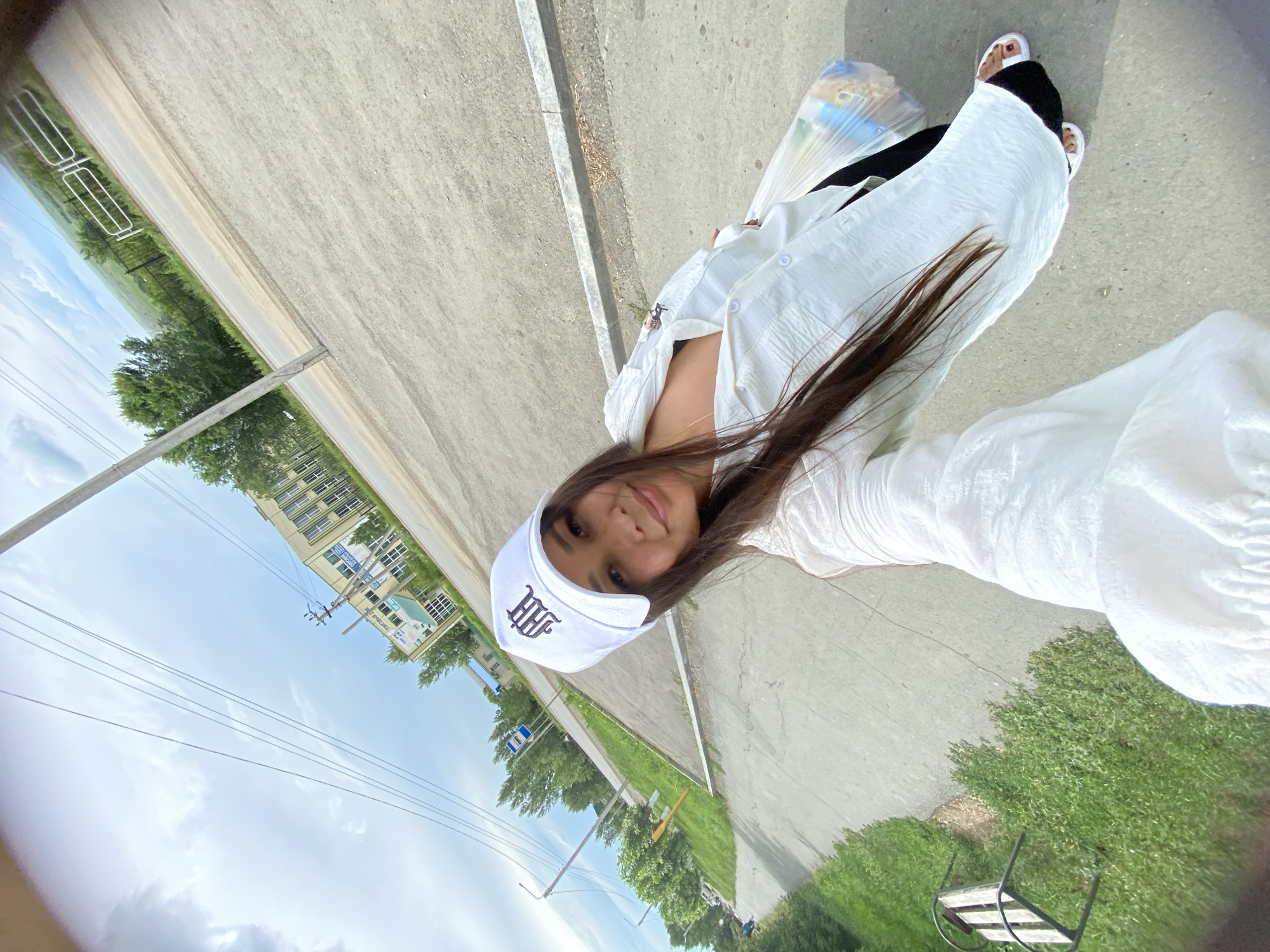 A young woman with long, dark hair is taking a selfie outdoors. She is wearing a white baseball cap with a dark embroidered logo, a white button-down shirt, and black pants with white sandals. She is holding a clear plastic bag filled with items. The background shows a paved road, a sidewalk, a grassy area with a bench, trees, a building with many windows, and a utility pole with wires. The sky is partly cloudy. The location is the city of Akyar, Russia.