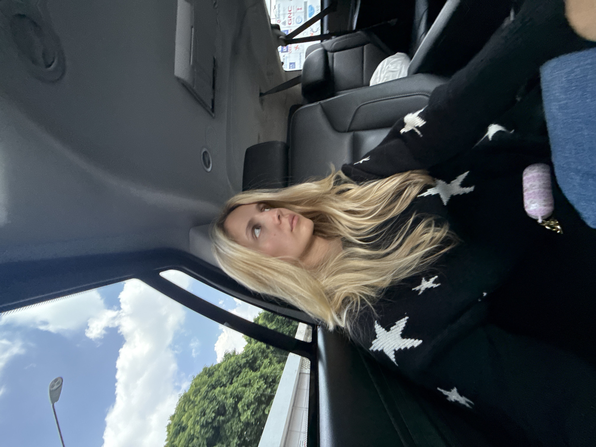 A blonde woman with long, wavy hair is seated in the back of a vehicle, looking out the window. She is wearing a black sweater with white stars. The interior of the vehicle is visible, with black leather seats and a sunroof. Outside, trees and buildings can be seen, as well as a street lamp against a blue sky with clouds. The scene appears to be in Mexico City, Mexico, based on the presence of a street lamp that is characteristic of the city's urban lighting.