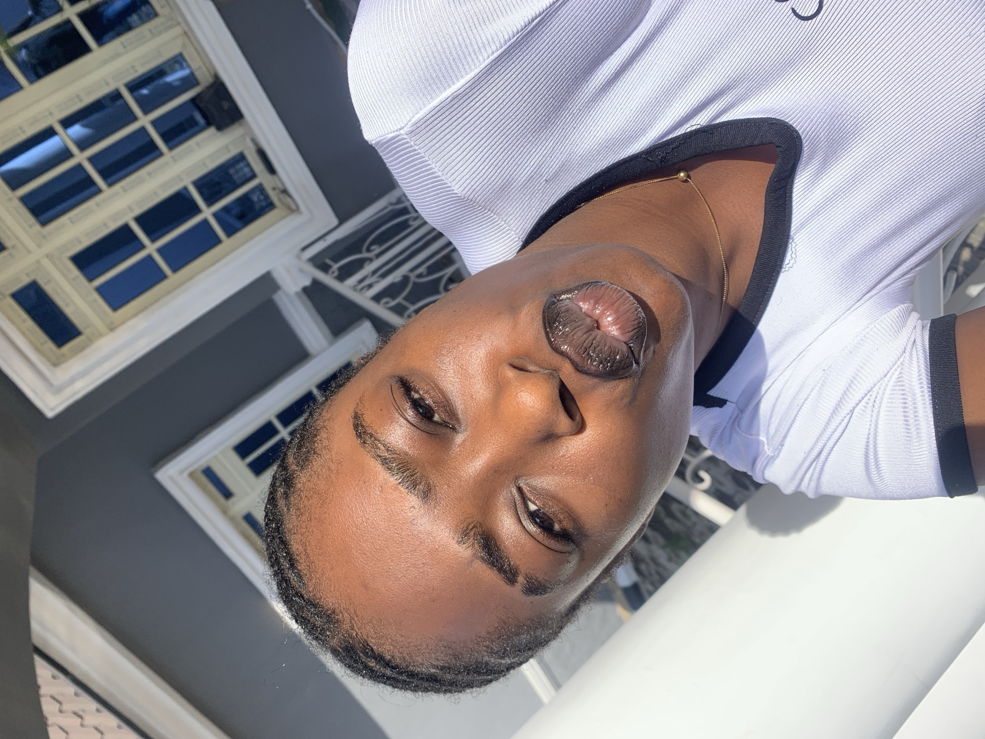 A young Black woman with braided hair and dark lipstick is taking a selfie outdoors in what appears to be a residential area of Gura Top, Nigeria. She is wearing a white ribbed t-shirt with black trim around the collar and a thin gold chain necklace. The background shows a gray building with white-trimmed windows and a white railing, indicating an urban or suburban setting. Sunlight casts shadows on her face and the white pillar to her left.