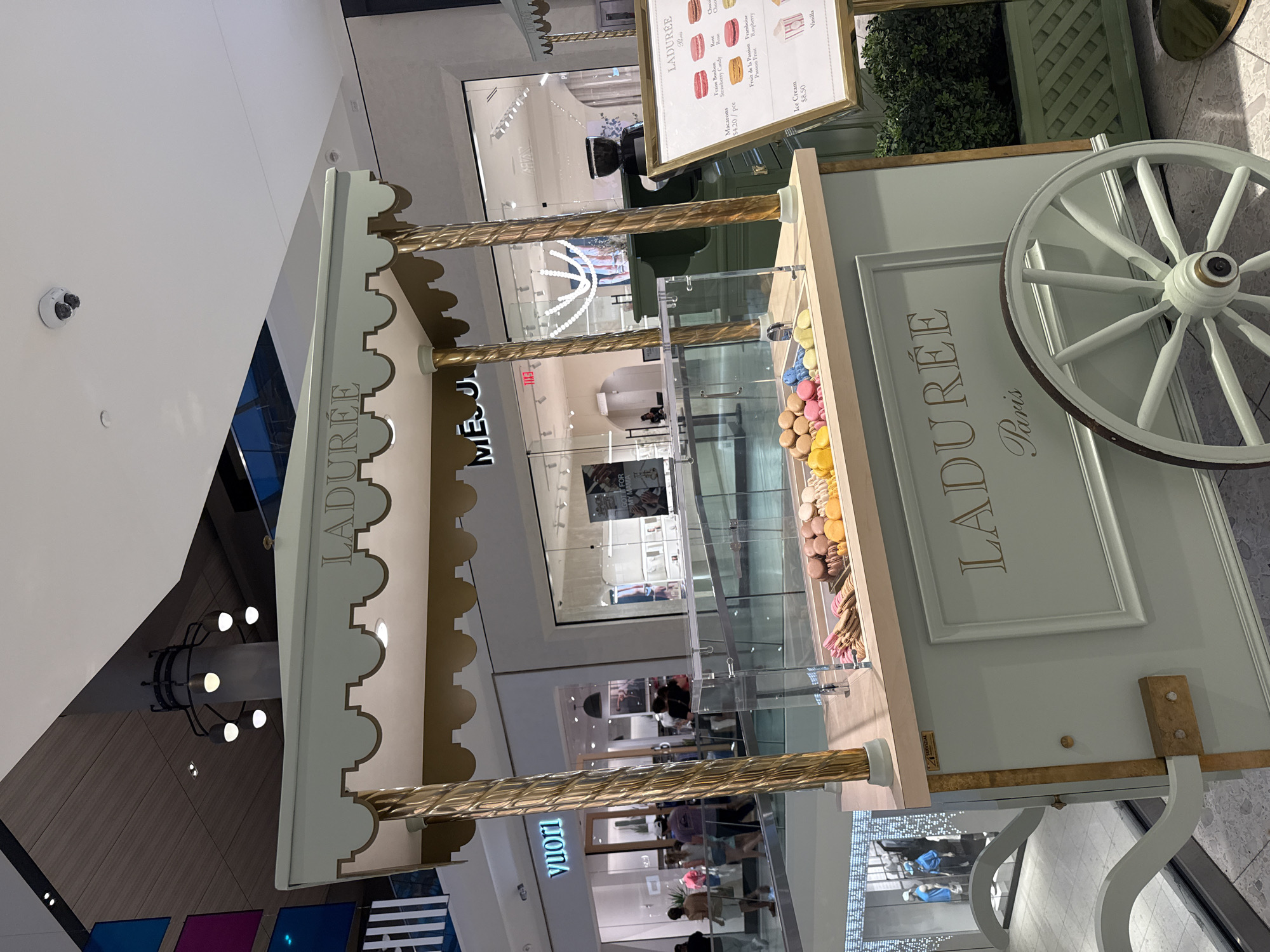 The image provides an overhead view of a bustling, multi-level shopping mall in Miami, United States of America, centered around a Ladurée macaron cart. The cart is styled in light mint green and gold, featuring ornate details like a scalloped canopy, twisted gold pillars, and a large decorative wheel. "LADURÉE Paris" is prominently displayed on the cart's side and canopy.
Inside a clear glass display case on the cart, a vibrant assortment of macarons in various colors, including blues, yellows, pinks, and browns, are neatly presented. An attached gold-framed menu board lists "Macarons $4.20/pce" with flavors such as Fraise Bonbon (Strawberry Candy), Rose, Chocolat, Fruit de la Passion, Framboise (Raspberry), and Vanilla. "Ice Cream $8.50" is also advertised. A small label on the cart reads "SERVIZIAL".
The background reveals multiple retail storefronts across different levels of the mall. Visible store names include "vuori" and a partial "MEJ" (likely "MES JUICES"), along with another sign displaying "GOT FOR". Planters with greenery are situated behind the cart. Numerous shoppers and mall visitors are seen moving throughout the bright, artificially lit environment. A white security camera is mounted on an upper white wall. The scene depicts a typical, active day in an upscale commercial center.