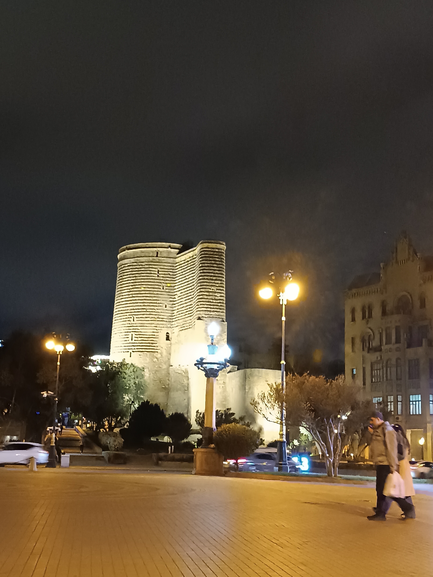 The image depicts the Maiden Tower in Baku, Azerbaijan, at night. The cylindrical tower, illuminated with horizontal bands, stands prominently against the dark sky. A section of the surrounding city wall is also visible. In the foreground, the paved square is illuminated by streetlights, creating an orange glow. A few people are walking on the right side of the image. Trees and bushes are scattered around the base of the tower. A tall building is partially visible on the right side of the image. White cars are parked on the left side of the image.