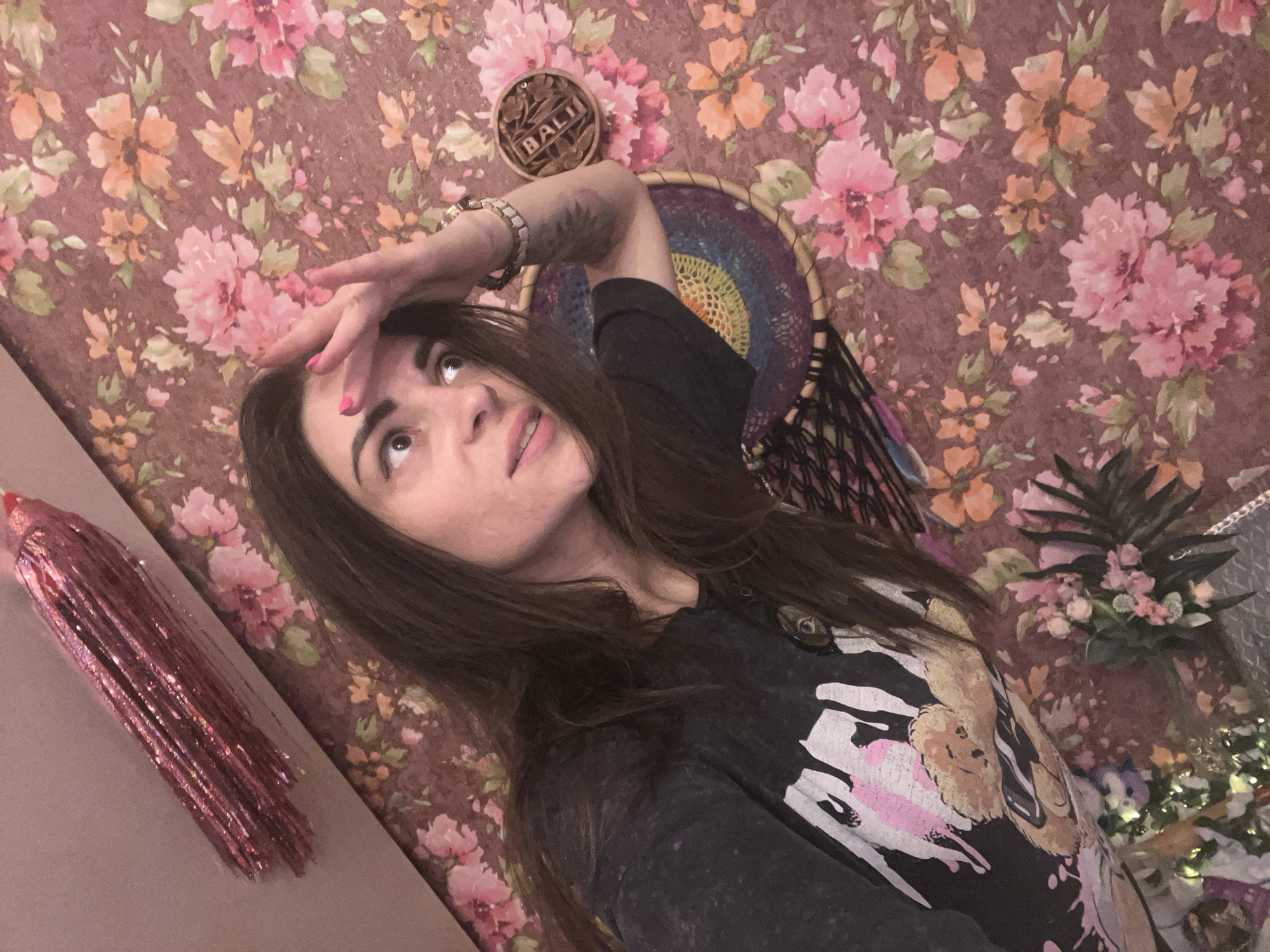 A woman with dark hair is taking a selfie in front of a floral wallpaper. She is wearing a dark t-shirt with a graphic print. Her right hand is raised to her forehead. To her left, a pink glitter tassel hangs diagonally. A dreamcatcher and a small wooden bowl with the word "BALI" are visible on the wall behind her. The image is taken from a high angle, looking down at the woman. The location is not specified in the provided information.