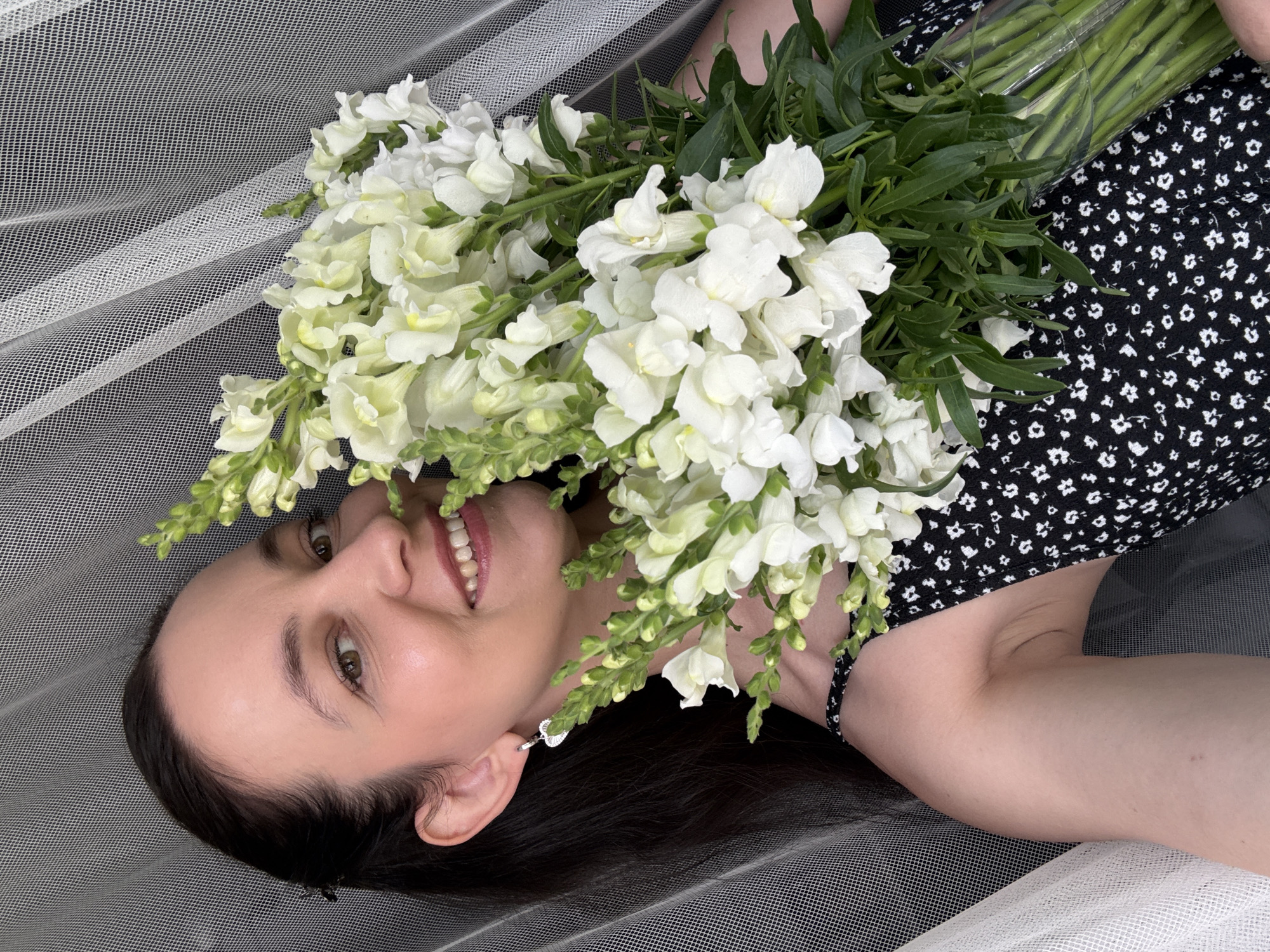 A young woman with dark hair and brown eyes smiles at the camera, holding a large bouquet of white snapdragons. She is wearing a black and white floral dress and a silver heart-shaped earring. The background is a sheer white curtain. The scene appears to be indoors.