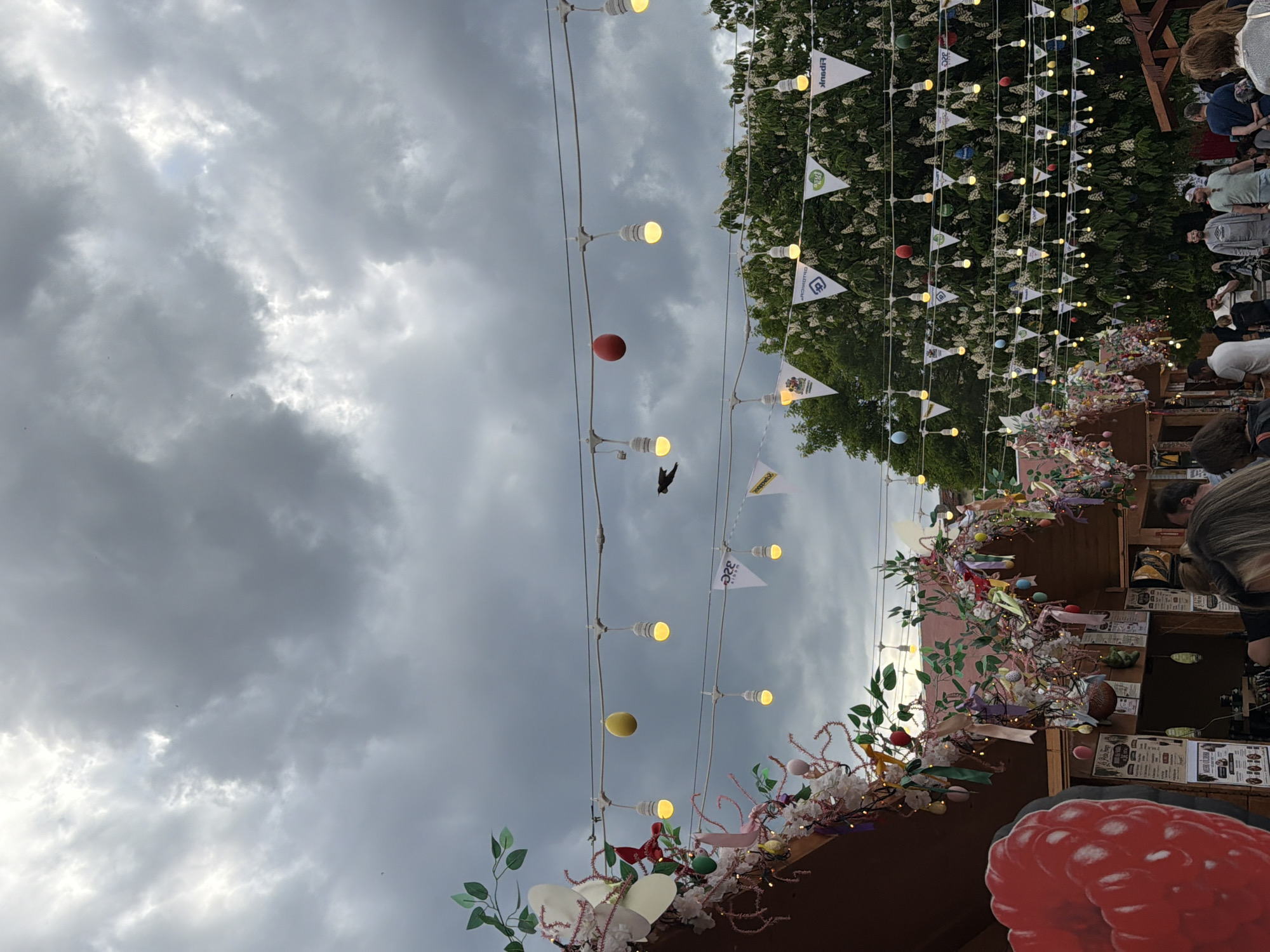 Captured from below, the photo presents a scene, possibly an open-air market, under an overcast sky. Strings of lights and pennant banners are strung overhead. Lower, stands are adorned with decorative elements like egg-shaped ornaments, foliage, and flowers. A large raspberry cutout is visible in the foreground. People are present in the background, with some appearing to be customers. The location may be Sofia, Bulgaria.