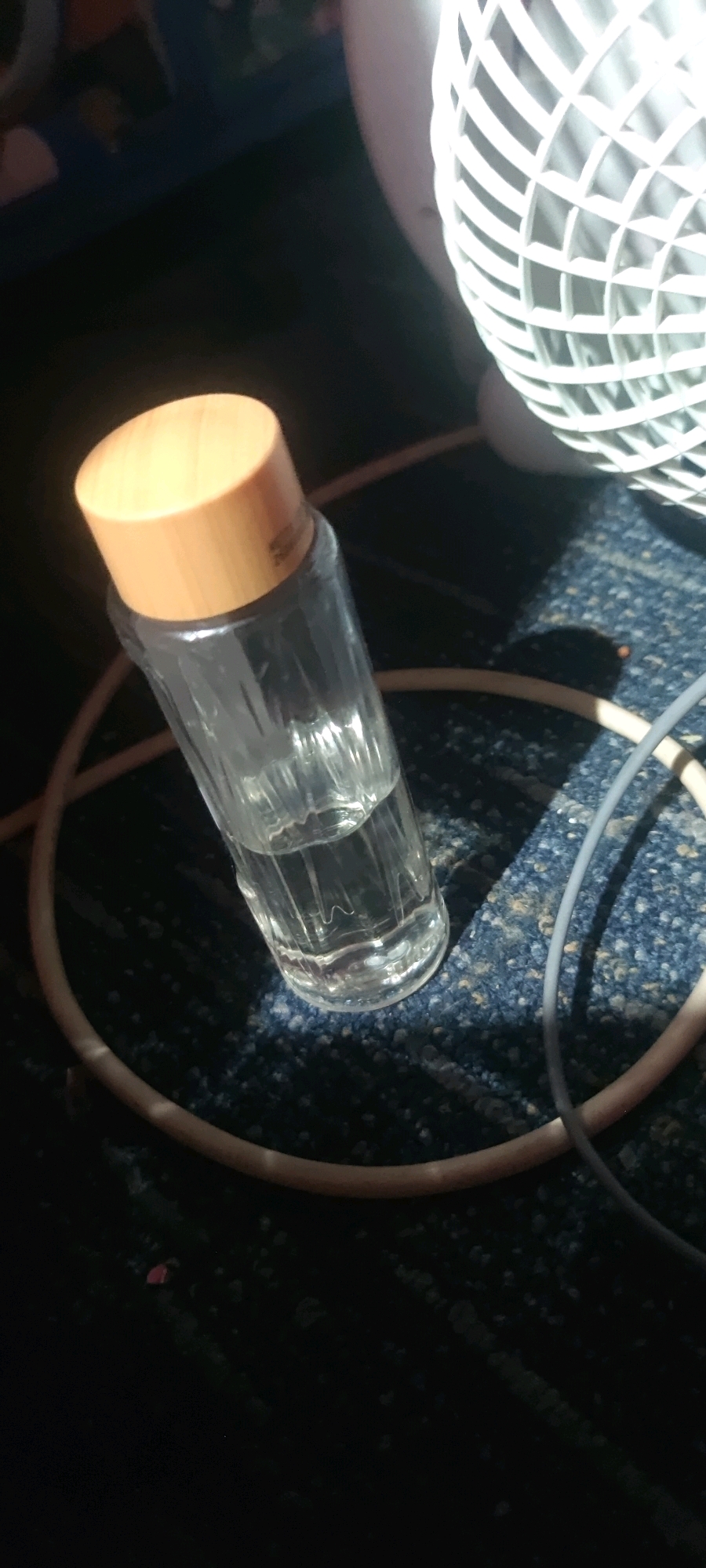 In the city of Gambarsari, Indonesia, a close-up, dimly lit photograph captures a glass bottle with a wooden lid, partially filled with a clear liquid. The bottle is positioned on a dark, textured surface, possibly carpet or fabric, which is blue and flecked with lighter particles. Two cables, one beige and one gray, are visible and coiled around the bottle, suggesting an indoor setting with electronic equipment. In the upper right corner of the frame, a portion of a white fan is visible, its grille showing a diamond pattern. The lighting casts shadows, adding to the subdued atmosphere. No people are present in the image.