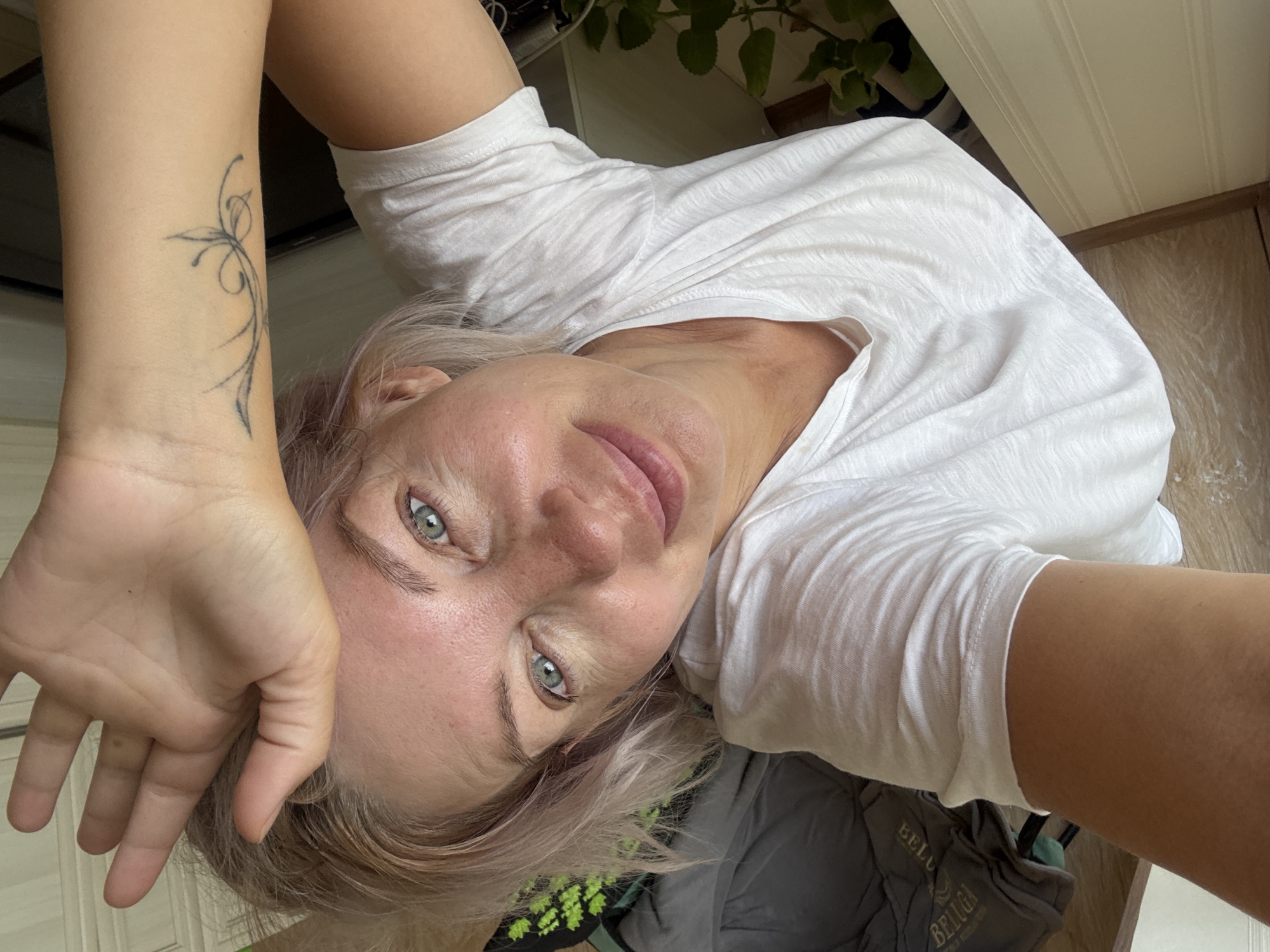 The image is a close-up, high-angle, selfie-style shot of a Caucasian woman. She has fair skin, light-colored hair that appears to be styled short and is slightly tousled. Her eyes are a striking blue-green. She is wearing a plain white t-shirt. Her right hand is raised and rests on her forehead, with her fingers splayed. A delicate, dark-ink tattoo, resembling stylized floral or vine elements, is visible on her left forearm. Her expression is relaxed, with a slight smile. The background is softly blurred, but hints of indoor elements are discernible. To the left, a dark gray comforter or blanket is visible, with some text in a lighter color, possibly a brand name like "BELUGA". Further left and slightly behind, there's a glimpse of what appears to be a green plant. The overall lighting suggests an indoor setting, possibly a bedroom or living area, with natural light illuminating the subject. The image does not contain any overt indicators of a specific location within the city of Sochi, Russia.