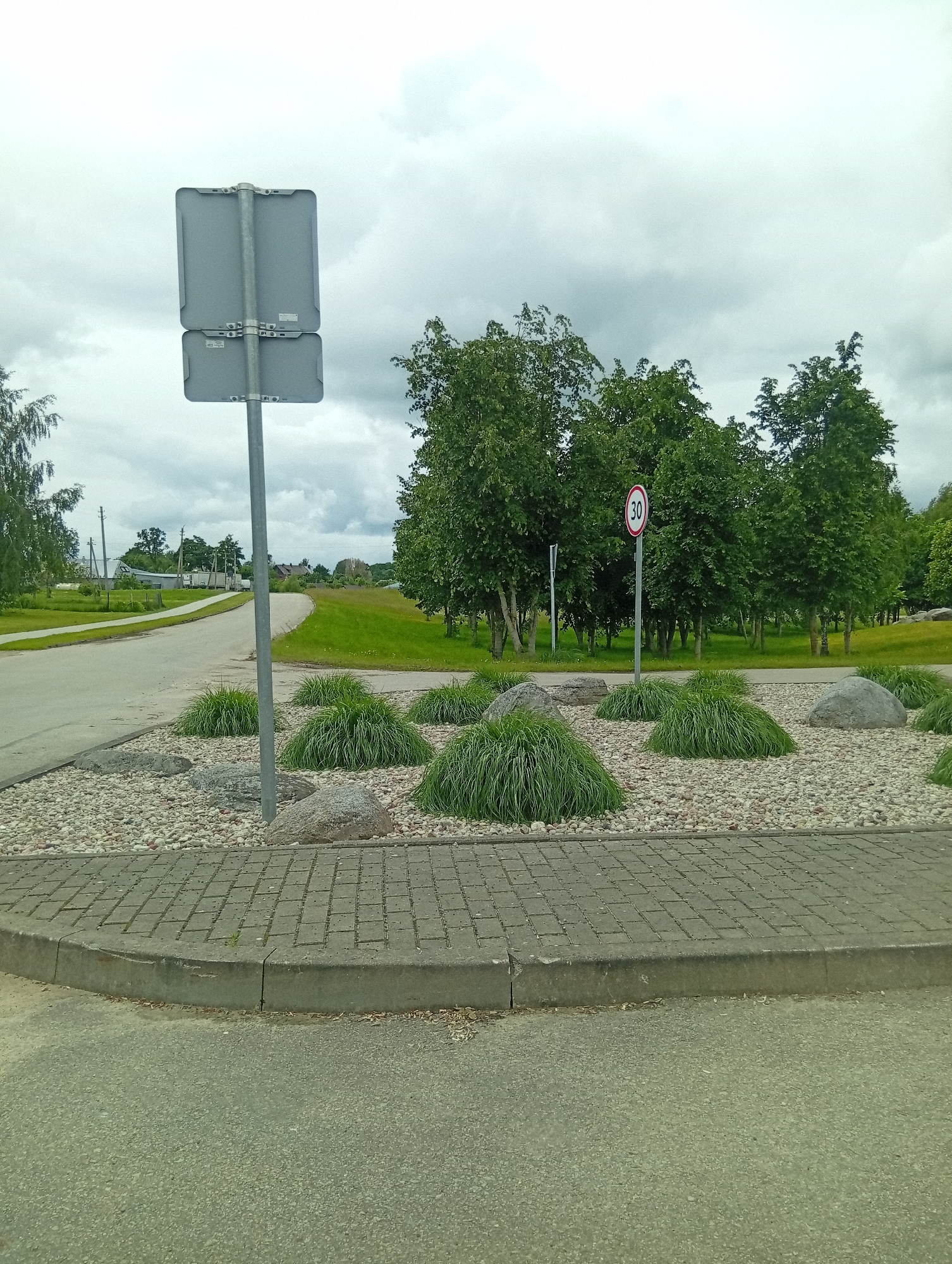 The image shows a road curving to the left, with a landscaped island on the right. The island features small, ornamental grasses, large rocks, and a bed of gravel. A speed limit sign indicating "30" is visible on the island. A grey traffic signpost with two rectangular signs is positioned on the left side of the image. In the background, there are trees, a green lawn, and some buildings. The sky is overcast. The scene appears to be in the town of Šaukėnai, Lithuania.
