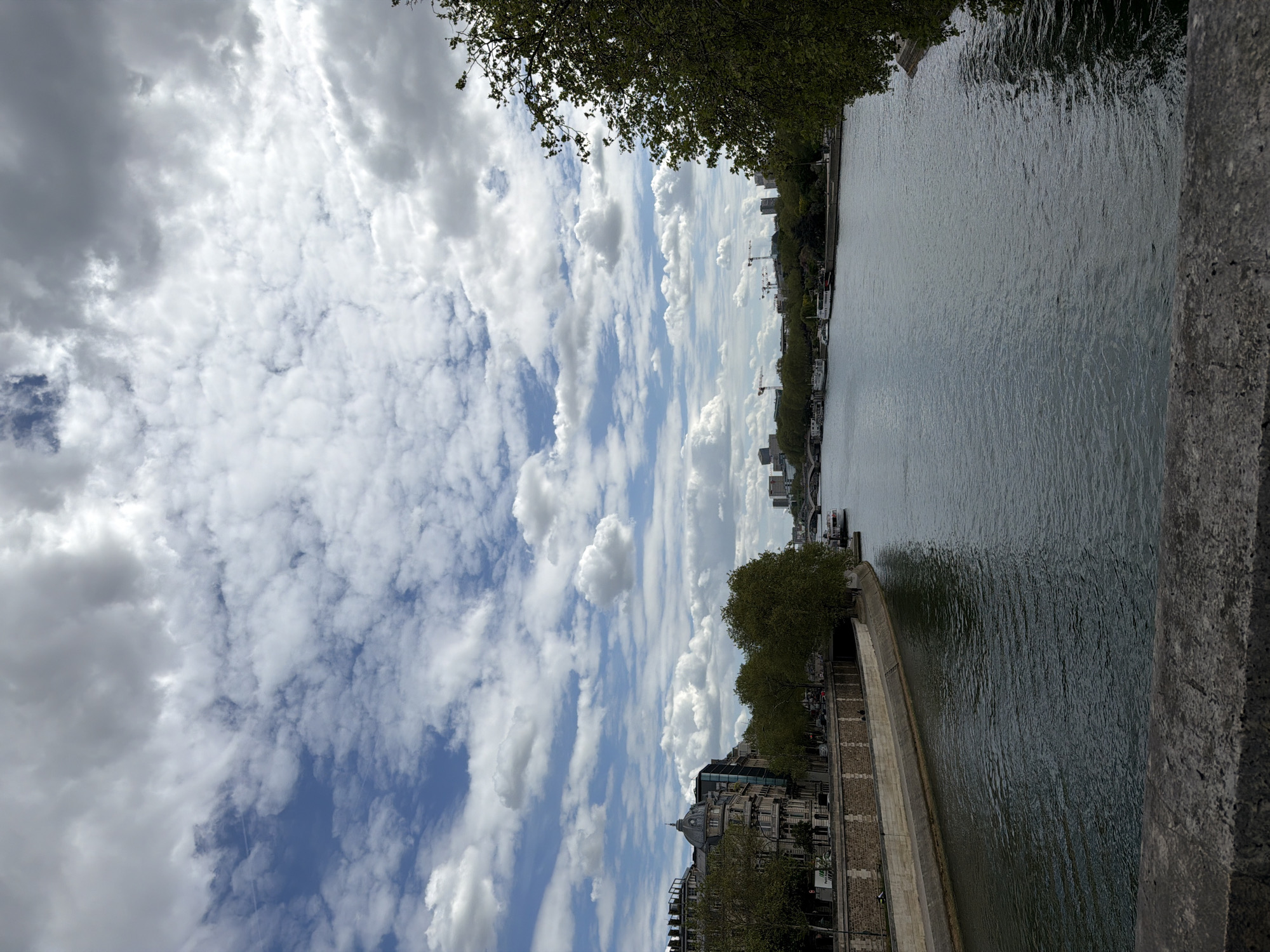 An outdoor, horizontal shot captures a view over a wide river, likely the Seine in Paris, France, under a partly cloudy sky. The perspective is from a slightly elevated position, possibly from a bridge or embankment, with a stone parapet visible on the right side of the foreground.
The sky occupies a significant portion of the upper frame, showcasing a dynamic interplay of fluffy white cumulus clouds against patches of bright blue. Some darker, more overcast cloud formations are also visible, suggesting changeable weather.
The river, a prominent subject, flows from left to right, its surface reflecting the cloudy sky and appearing a muted grey-green. Numerous small ripples are visible across the water, indicating slight movement or a gentle breeze.
Along the far bank, a dense cityscape stretches into the distance. Various buildings, some appearing older with classical architecture and others more modern, line the river. Several tall construction cranes rise above the skyline, signifying ongoing urban development. Green trees form a verdant line along this bank, partially obscuring the buildings.
The near bank, visible on the left side of the river, features a well-constructed stone embankment or quai, with more trees and buildings behind it. A building with a distinctive domed roof is visible among the structures on this side. What appear to be boats or barges are docked along this bank.
The time of day appears to be daytime, with natural light illuminating the scene. No specific human activity or identifiable text is clearly visible. The overall mood is calm and contemplative, characteristic of a scenic urban river landscape.