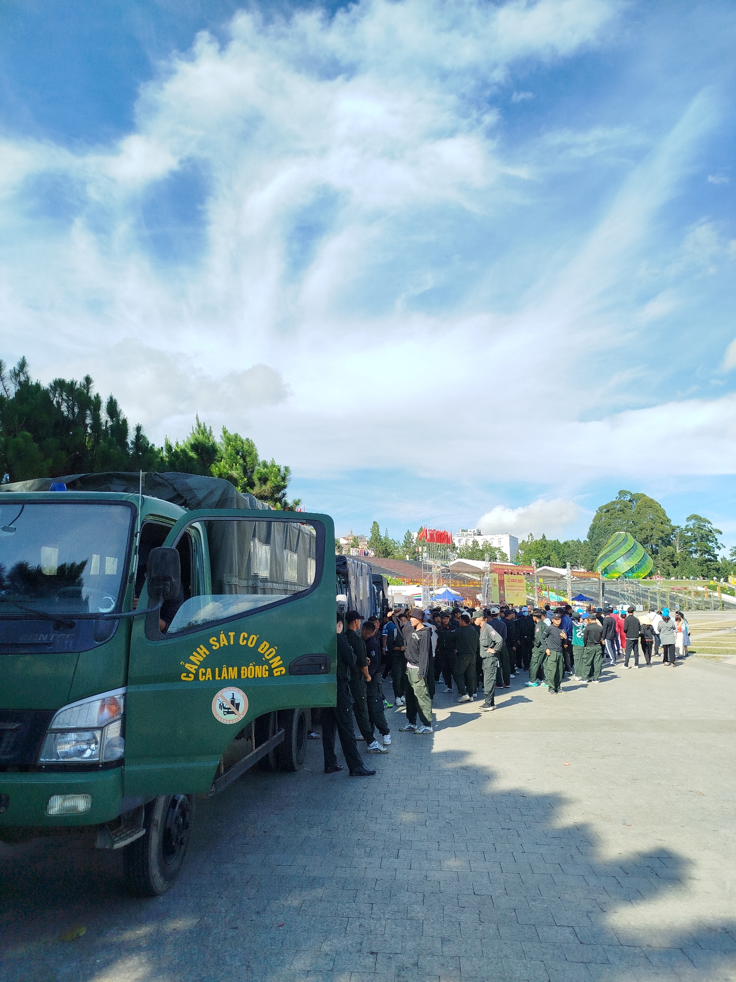 In the city of Da Lat, Vietnam, a group of uniformed individuals, likely law enforcement or security personnel, are gathered outdoors on a paved area, possibly for an event or a deployment. A green truck with "CẢNH SÁT CƠ ĐỘNG CA LÂM ĐỒNG" (Mobile Police Lam Dong) written in yellow on its side is parked on the left. The truck's passenger door is open, and its cargo area is covered by a dark tarp. A circular decal with an image of a crossed-out cigarette and alcohol bottle is affixed to the truck's door. The individuals are standing in a line, some facing the truck and others looking away. They are wearing dark uniforms and appear to be waiting or receiving instructions. In the background, there are trees, buildings, and a large, green, striped inflatable structure that resembles a hot air balloon. The sky is bright blue with scattered white clouds, suggesting a clear day. The ground is paved with grey interlocking bricks. The scene suggests an organized gathering, possibly related to a public event or security operation.