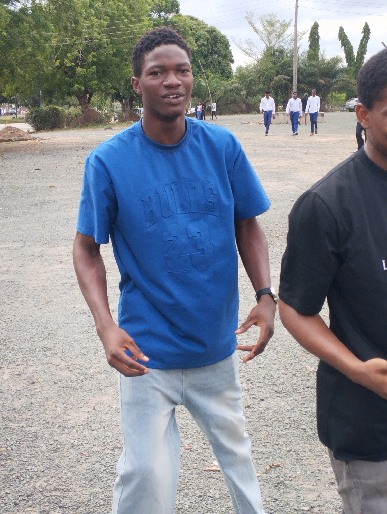 The image depicts an outdoor scene in Ihiagwa, Nigeria, characterized by an unpaved, gravelly surface under an overcast sky. In the foreground, a young adult male with dark skin and short, dark, curly hair is positioned slightly left of center, looking towards the viewer with a slight open-mouthed expression. He wears a royal blue short-sleeved t-shirt with "BULLS 23" embossed on the chest, light wash blue jeans, and a black digital watch on his left wrist. His hands are open and slightly extended at waist level. To his immediate right, partially visible, is another individual with dark skin wearing a black short-sleeved t-shirt with light lettering (partially visible "L") and grey trousers.
In the mid-ground, three young adult males walk from left to right across the gravel surface. All three are uniformly dressed in white long-sleeved button-up shirts and dark blue trousers. Further in the background, to the left, more indistinct figures are present. The background features lush green foliage from numerous trees, including large deciduous trees on the left and taller, slender trees, possibly palms, on the right. A utility pole stands vertically in the mid-ground, slightly right of center, with faint power lines visible overhead. A light-colored vehicle is partially visible in the far background to the right, and another dark vehicle is partially visible to the far left. The ground is predominantly light grey crushed stone or gravel with scattered darker patches.