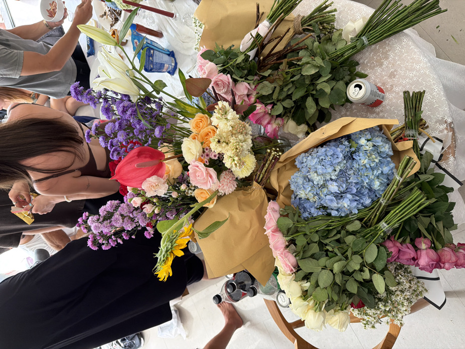 The image captures a vibrant, high-angle view of a bustling scene centered around a large assortment of fresh flowers, likely in preparation for an event or arrangement. The setting appears to be indoors, with bright, even lighting illuminating a white-covered table or work surface.
Numerous bundles and loose cut flowers dominate the frame, showcasing a wide variety of colors and species. Prominently featured are large clusters of light blue hydrangeas, still partly encased in brown kraft paper, alongside several bundles of roses in shades of white, light pink, and deeper pink/purple. Other flowers include striking white lilies, vibrant purple asters or similar small purple blooms, a bold red anthurium, clusters of orange and yellow roses, creamy white/light yellow textured flowers, and a distinct yellow sunflower head. Many of the flower bundles are neatly tied with string or rubber bands, and some are wrapped in protective brown paper. Small white filler flowers are also visible among the larger blooms.
Several individuals are partially visible, engaged in the activity. In the upper left, a person wearing a grey t-shirt holds a small white bowl containing what appear to be brown, irregularly shaped pieces. Below them, closer to the center-left, another person with long dark hair, wearing a dark brown top, is seen leaning over the flowers, possibly holding a tool like scissors or clippers, suggesting they are actively preparing or arranging the blooms. This person has a tattoo on their arm. In the bottom right corner, a bare foot and part of a leg wearing dark pants are visible, indicating another person standing nearby.
The white surface beneath the flowers has a delicate, textured pattern, possibly a lace tablecloth. Amidst the flowers, an open silver aluminum can with red markings is visible near the top right. On a wooden surface in the lower right, beneath some of the flowers, several bottles and cans are present, including a bottle that appears to be red wine and other aluminum cans. In the background, on the white surface, a blue package, faintly identifiable as "OREO," is also visible.
The overall impression is one of a busy and creative environment, possibly a floristry workshop, a wedding preparation, or a large flower delivery being processed. The abundance and variety of fresh flowers create a colorful and lively atmosphere.