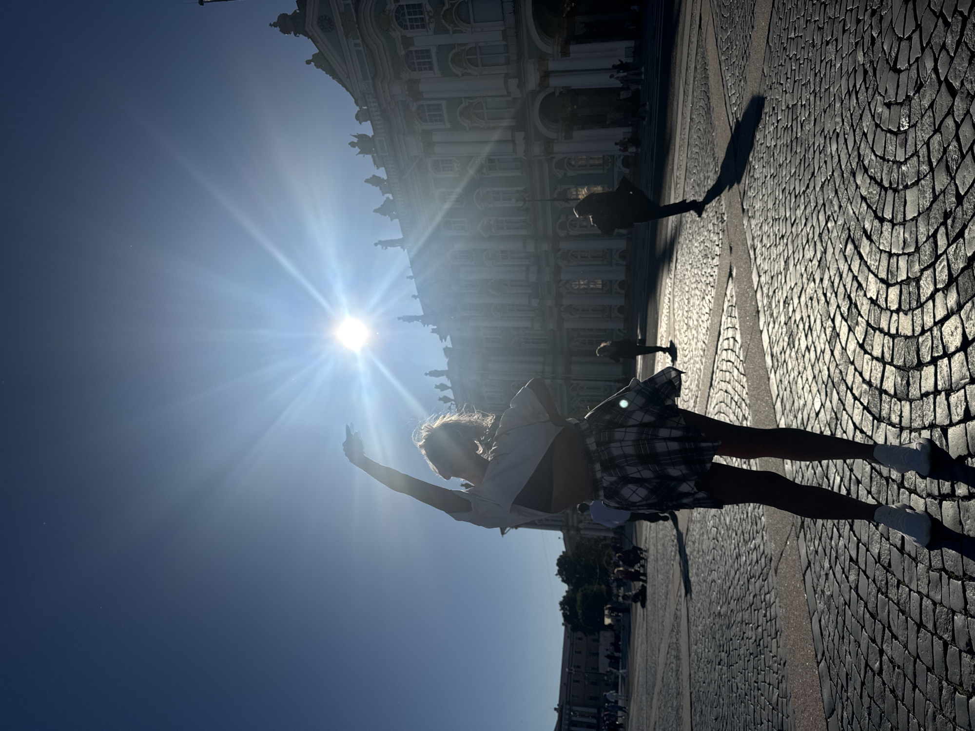 In Saint Petersburg, Russia, a young woman is silhouetted against a bright sun in Palace Square, near the Hermitage Museum. She is wearing a white t-shirt, a plaid skirt, and white sneakers. Her arms are raised, and her body is angled as if she is dancing or posing. The sun is high in the sky, creating a lens flare and casting long shadows across the cobblestone plaza. Other people are visible in the background, some walking and others sitting, but they are out of focus. The architecture of the Hermitage Museum, with its ornate facade and statues, is visible behind the woman. The scene is bathed in the warm light of a sunny day.