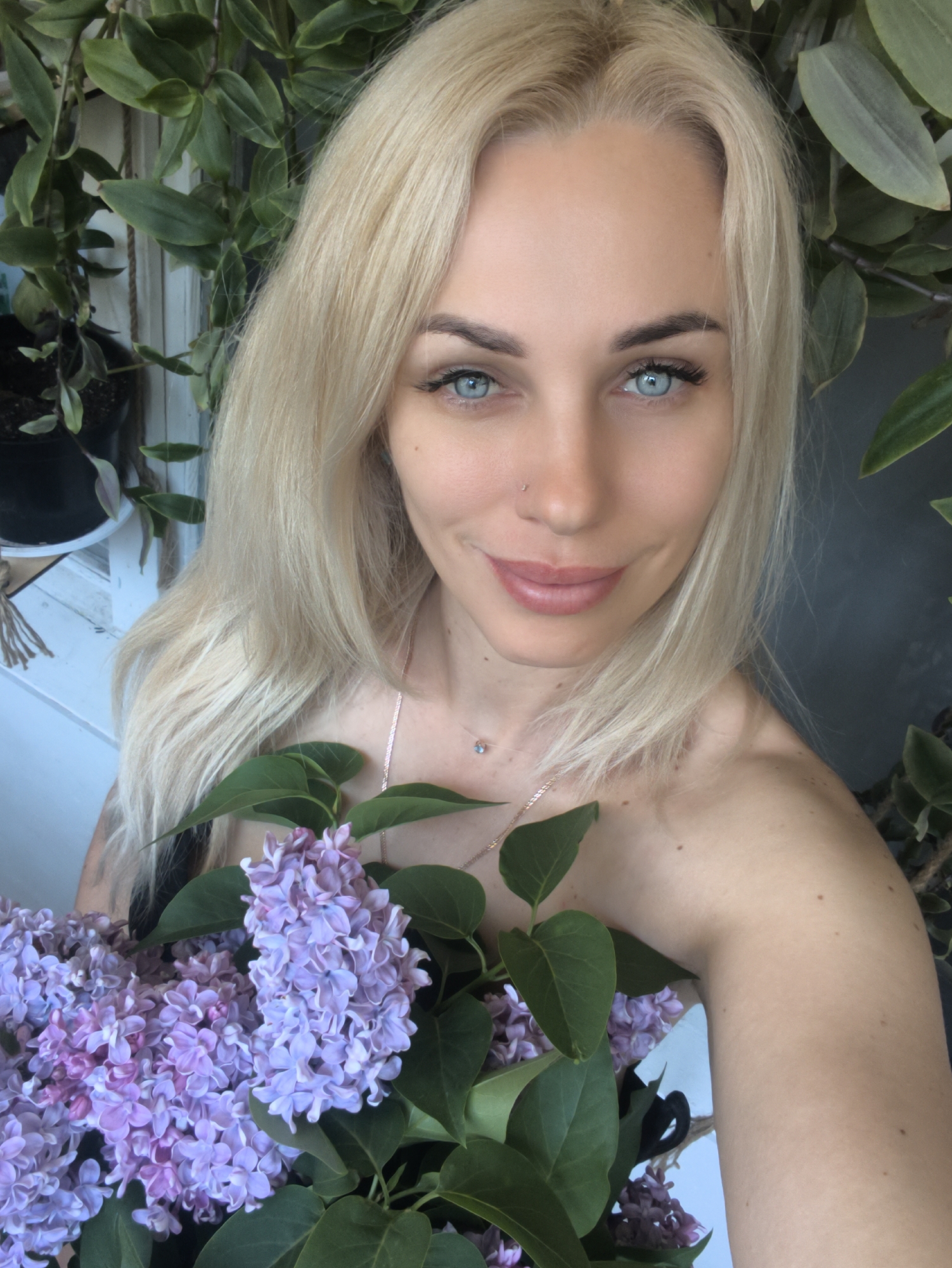 A woman with fair skin, blonde hair, and blue eyes is holding a bouquet of purple lilacs. She is wearing a delicate necklace and a nose ring. The background consists of leafy green plants and a white wooden structure. The location and city are not discernible from the image.