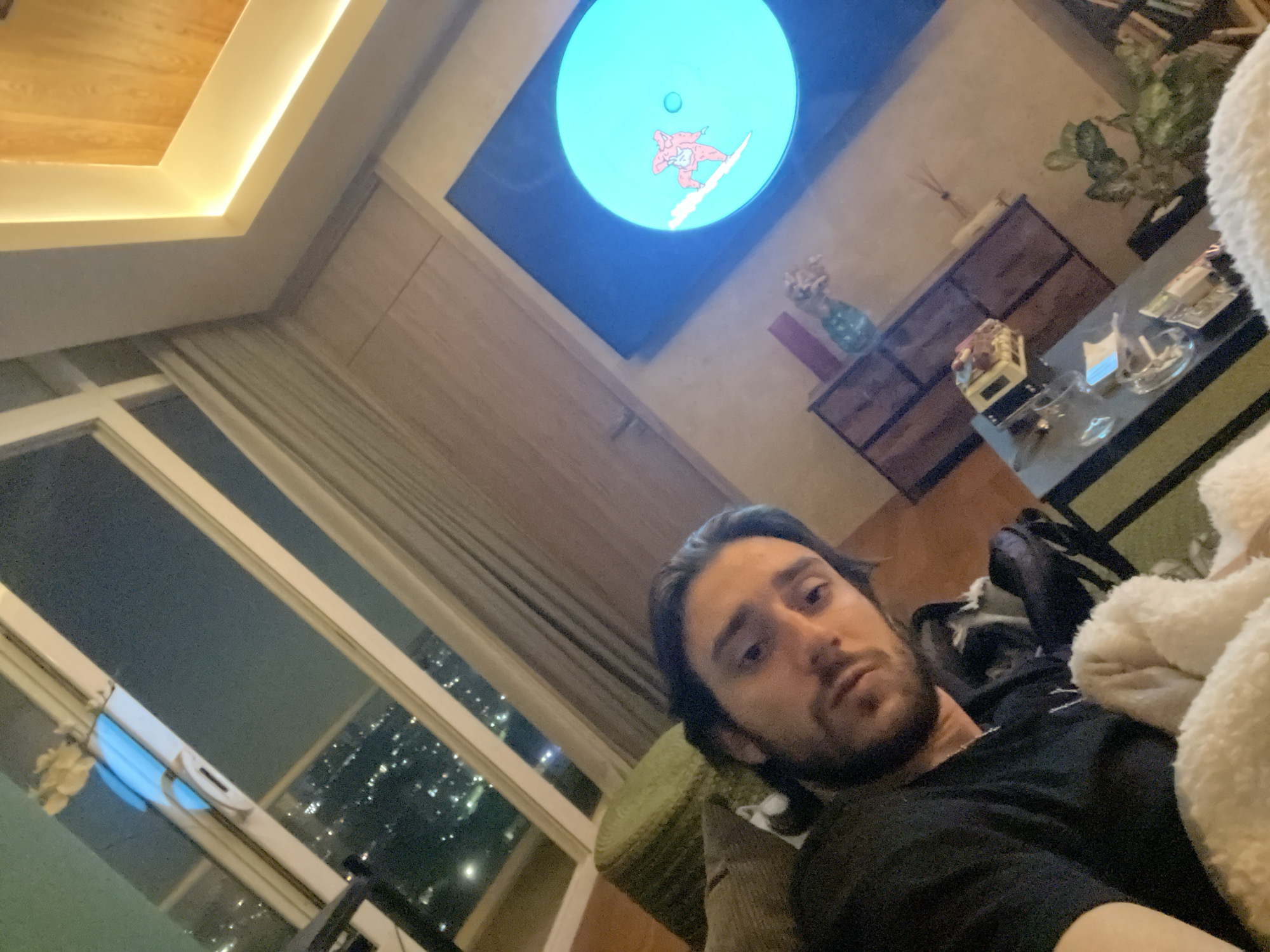 The photograph captures a man with dark hair and a beard, wearing a black shirt, seemingly relaxing indoors. He appears to be reclining on a couch or chair, partially covered by a fluffy white blanket. In the background, a window reveals a cityscape at night, the lights of which suggest a dense urban area. The location can be identified as Huixquilucan de Degollado, Mexico, based on the urban sprawl visible through the window. A television screen mounted on the wall displays a cartoon figure on a snowboard. The lighting in the room is subdued, with a warm glow emanating from the ceiling. Other elements in the scene include a coffee table, various decorative items, and plants.