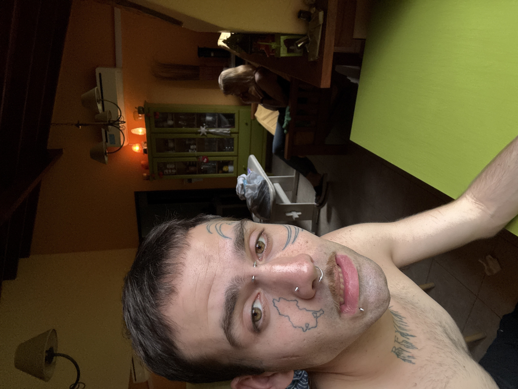 A selfie depicts a shirtless person with facial tattoos and piercings. The tattoos include geometric shapes on the forehead and a map outline on the cheek. The person has a nose ring and a labret piercing. In the background, a woman is seated at a table with a green surface. Behind her is a green cabinet with glass doors. A high chair is also visible. The room appears to be indoors, with artificial lighting. It is not possible to determine if the location is Villa General Belgrano, Argentina, solely from this image.