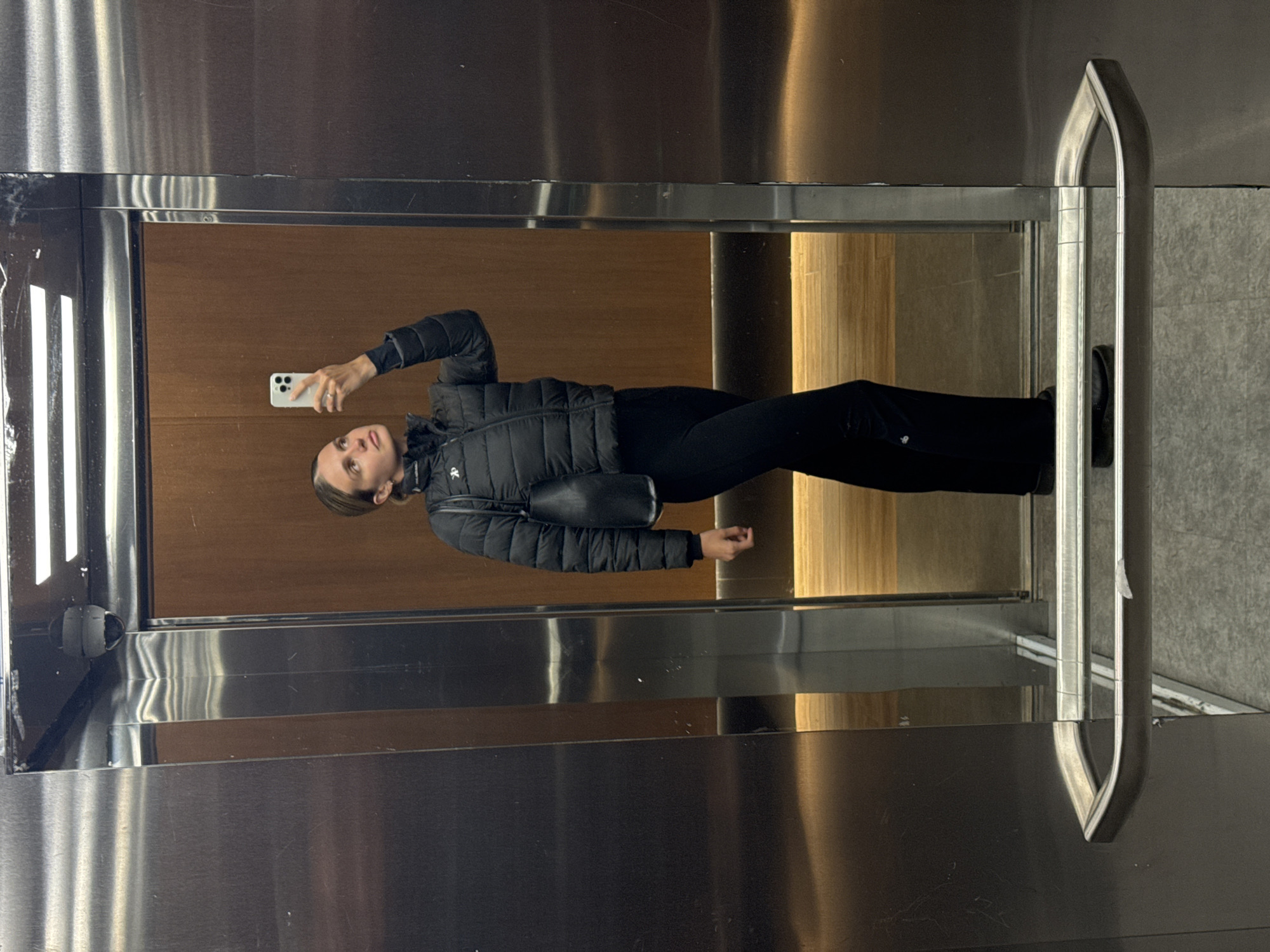 The image shows a woman taking a mirror selfie inside what appears to be an elevator or a room with mirrored walls. The primary subject is the woman, reflected prominently in the mirror. She is dressed in a black puffer jacket, black trousers, and has a black cross-body bag. Her dark hair is pulled back, and she is holding a white smartphone, likely an iPhone, in her right hand, pointing it towards her reflection. Her expression is neutral as she looks towards the phone.
The setting is characterized by a reflective surface that mimics a large mirror, creating the illusion of depth. The background of her reflection consists of warm, vertical wooden paneling. On the left side of the reflection, there are bright, vertical strip lights. To the right of her in the reflection, a metallic handrail is visible, which also appears to be part of the actual elevator or room structure. The walls surrounding the mirrored area are made of a brushed metallic material, possibly stainless steel, which also reflects some light. The floor, partially visible near the handrail, appears to be a light grey tiled surface. A small, indistinct logo, possibly "CK," is visible on the left side of her jacket. No other discernible text is visible. The lighting is artificial, suggesting an indoor setting.