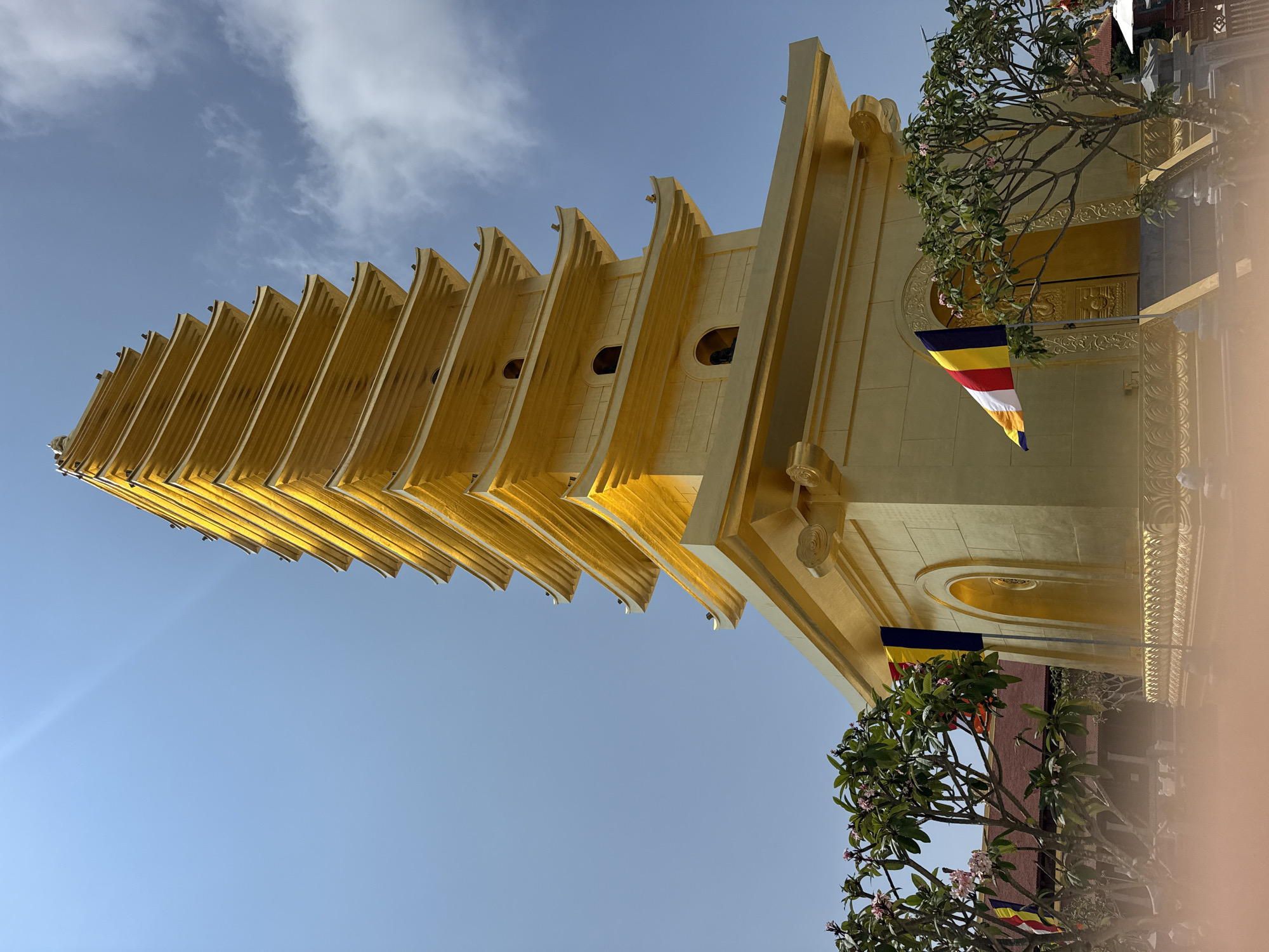A low-angle, outdoor photograph captures a multi-tiered golden tower or structure against a light blue sky with few clouds. The structure has multiple receding tiers, reminiscent of a pagoda, and is adorned with Buddhist flags. The structure is in Diên Khánh, Vietnam. Trees with green leaves and pink flowers are visible near the base of the structure. A person is out of focus on the right.