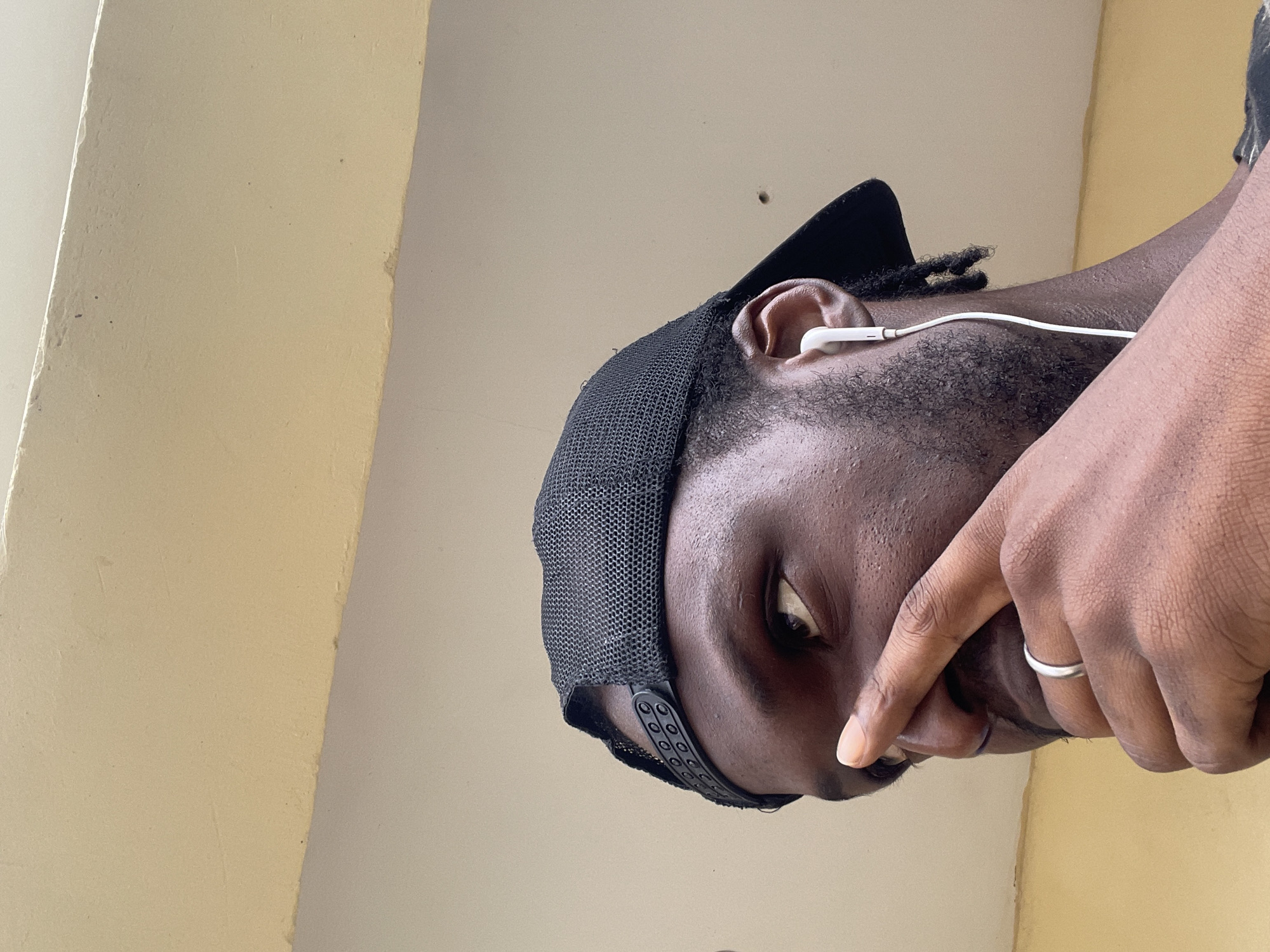 The image shows a close-up, low-angle shot of a Black person wearing a black baseball cap and white earbuds. Their right hand is raised, with a finger touching their nose, and a silver ring is visible on their left ring finger. The person is looking to the left, with their eye clearly visible. The background consists of two distinct sections: the upper portion is a light cream-colored wall, and the lower portion is a similar cream color but with a rougher texture. A very faint shadow is visible on the wall in the upper right quadrant. The scene appears to be outdoors, possibly against a building. There is no indication of the specific location within Abuja, Nigeria.