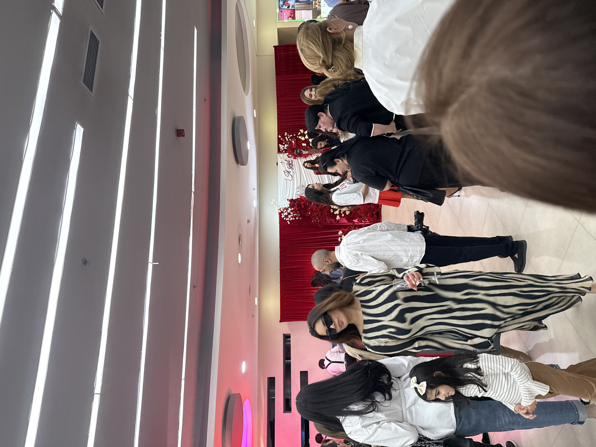 A group of people are gathered at what appears to be a celebration, potentially a birthday, in Yerevan, Armenia. The scene is indoors, indicated by the bright artificial lighting, long lines of ceiling lights, and walls. There is a decorative setup with red drapery, flowers, and signage visible in the background, suggesting a stage or focal point for the event. Attendees are dressed in a variety of outfits, including formal dresses, casual attire, and shirts, including a black and white zebra-striped dress. Several people are standing and appear to be watching or participating in the event. A woman in sunglasses holds a wine glass. A small child is wearing a white shirt with black stripes and a large white bow in her hair. The floor is tiled.