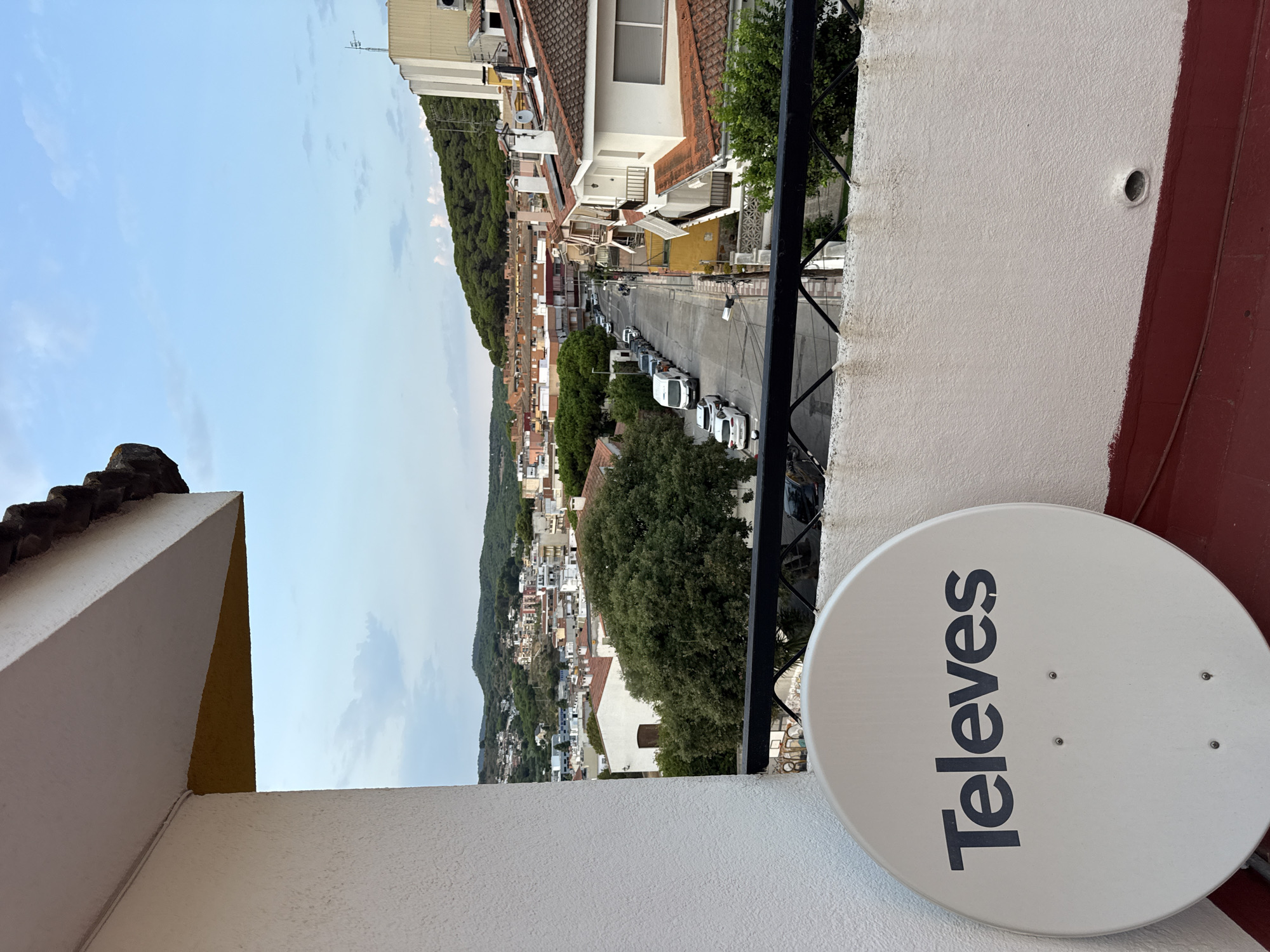 The image depicts a view from a balcony in Blanes, Spain, overlooking a residential street lined with buildings. In the foreground, a white satellite dish with the brand "Televes" is visible. The balcony railing is black and metal. Beyond the balcony, the street curves to the right, with parked cars and trees on either side. Buildings with red-tiled roofs and white or cream-colored facades are interspersed with lush green trees. In the background, the town extends up a hillside covered in more buildings and dense vegetation, leading to a tree-covered mountain under a partly cloudy sky. The scene appears to be captured during the late afternoon or early evening. No people are visible.