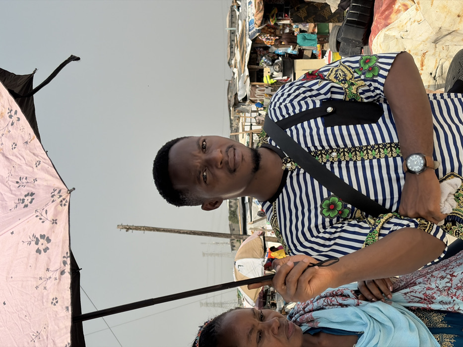 The image depicts an outdoor scene in what appears to be a market or informal settlement in Igboefon, Nigeria, under an overcast or hazy sky.
**Individuals:**
1. **Primary Male Subject:** Positioned slightly right of center, a young adult male of Black ethnicity looks directly at the camera with a neutral expression. He has short, dark hair and a short beard. He wears a short-sleeved collared shirt with horizontal blue and white stripes, adorned with embroidered green and red floral patterns on the chest and sleeves, and a black pocket with a silver stud on the left chest. A black shoulder bag strap crosses his chest. On his left wrist, he wears a dark-faced wristwatch with a brown leather strap. His right hand holds the dark handle of an umbrella, while his left hand holds a light-colored folded cloth at waist level.
2. **Secondary Female Subject:** Partially visible to the left and slightly behind the male subject, an adult female of Black ethnicity is present. Only her head and upper torso are visible. She appears to be looking downwards or away from the camera, displaying a somber expression. Her dark hair shows colorful beadwork on top. She is wearing a light blue head covering or shawl, with an additional patterned fabric (red/brown floral design on a dark background) draped over her shoulder.
**Objects and Environment:**
* **Umbrella:** A large, open, light pink umbrella with a dark floral pattern occupies the top portion of the frame, providing shade. Its dark handle is held by the primary male subject.
* **Background Structures:** The background consists of numerous low-rise, rudimentary structures, indicative of market stalls or makeshift dwellings. These structures feature corrugated metal roofs and various goods displayed, including what appear to be stacks of tires.
* **Utility Pole:** A wooden utility pole stands upright behind the primary male subject, slightly to his left.
* **Power Infrastructure:** In the distant background, what appear to be power lines and transmission towers are visible against the hazy sky.
* **Other Individuals:** A person wearing light blue shorts is visible in the background, near one of the market stalls, indicating activity.
* **Ground:** The ground level is largely obscured by subjects and background elements, but visible areas suggest unpaved earth or dirt.