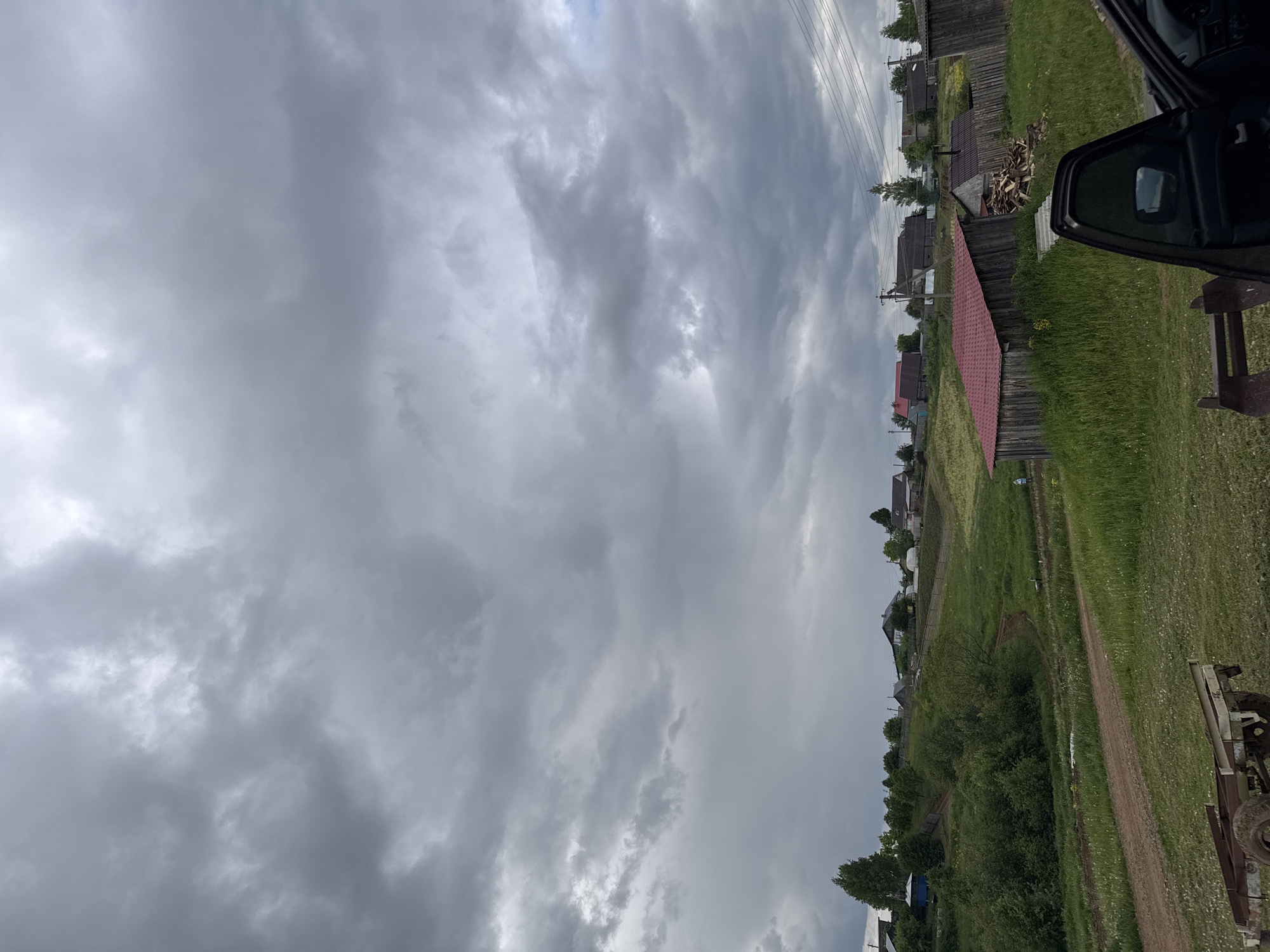 This media file captures a rural landscape under a heavily overcast sky, likely set in Бородулино, Russia. The dominant feature is the sky, which fills over half of the frame with dense, grey clouds, suggesting dull, diffused light and potentially impending rain.
The lower portion of the image depicts a village scene. On the right side, the open black door of a car is visible in the immediate foreground, indicating the perspective of the shot. Alongside it, a rustic wooden bench or structure is partially seen, and the ground is covered in vibrant green grass.
Further into the midground and background, a line of wooden houses stretches across the right half of the image. These structures vary, with some featuring older wooden fences. One house stands out with a distinct red roof, and a pile of chopped firewood is stacked nearby. Utility poles with multiple overhead wires run parallel to the houses, receding into the distance.
A dirt path or road meanders from the bottom center of the frame, curving slightly rightwards and leading through the village landscape. The surrounding fields are covered in lush green grass, interspersed with small wildflowers. Various trees and bushes are scattered throughout the scene, particularly along the distant horizon and bordering the path. Part of what appears to be agricultural equipment or a trailer is visible in the bottom right corner.
No specific human or animal activity is depicted. The time of day appears to be daytime, though the heavy cloud cover prevents a more precise determination. There is no visible text within the image. The overall impression is one of a quiet, tranquil rural setting.