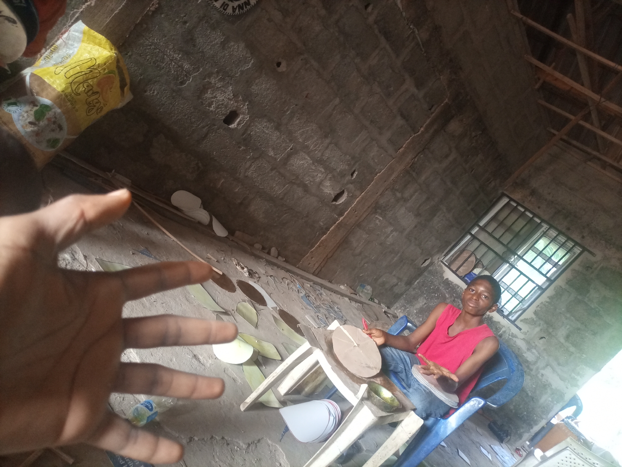 An outdoor, low-angle shot shows a young person sitting on a blue plastic chair in an unfinished concrete building. The person, wearing a pink tank top and blue jeans, is sitting at a low table with various craft materials. There are round paper cutouts and a wooden disk on the table. The person is holding a red object, possibly a pen or marker, and is gesturing with their left hand. The building has a concrete floor and walls, with exposed wooden beams on the ceiling and a window with metal bars. In the foreground, a blurred hand is visible, as if the photographer is in the frame. Other objects in the scene include a yellow bag with writing on it and a plastic water bottle.