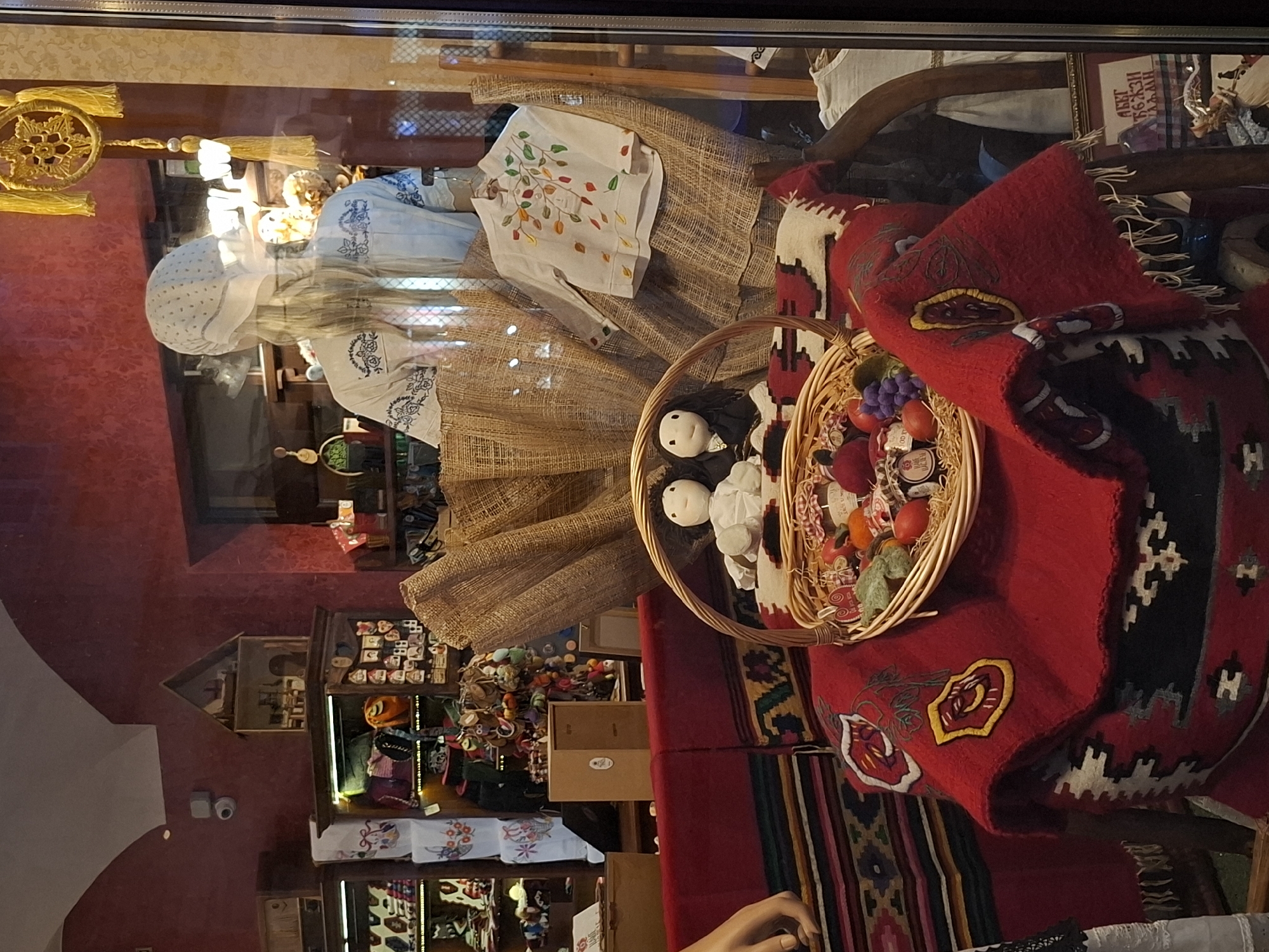 The image depicts a shop window display, likely in Novi Sad, Serbia, filled with a variety of traditional and folk-art items.
The central element is a mannequin dressed in what appears to be traditional Serbian clothing. The mannequin has a white headscarf, a blouse with embroidered designs, and is draped in a textured woven material.
A woven basket filled with dolls and small decorative items sits in front of the mannequin. A red, patterned rug or textile is draped over the display.
The shop's interior is visible through the window. Shelves are stocked with various items, including clothing, souvenirs, and crafts. The walls of the shop are a deep red color. A surveillance camera is visible on the wall.
