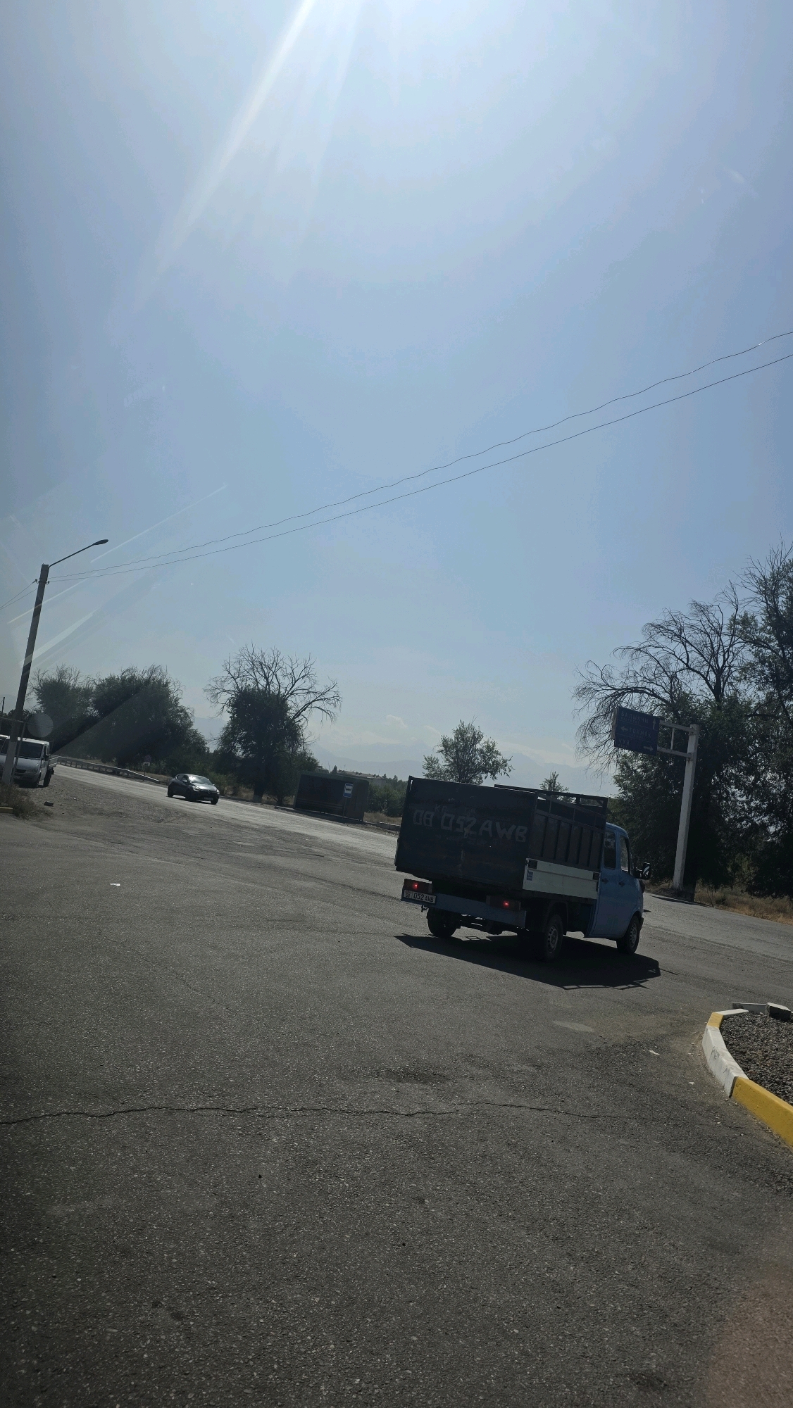 The image shows a light blue truck driving on a paved road. The truck has a dark-colored bed with white lettering that appears to read "08 052 AW8". A license plate on the rear of the truck reads "08 052 AW8". The truck is in motion, and its shadow is cast on the road behind it. The road is cracked in several places. In the background, there are trees, some of which appear to be bare. There are also mountains visible in the distance under a clear blue sky with bright sunlight. A street light is visible on the left side of the image. The scene appears to be an intersection or a curve in the road, with a yellow and white curb on the right side of the image.