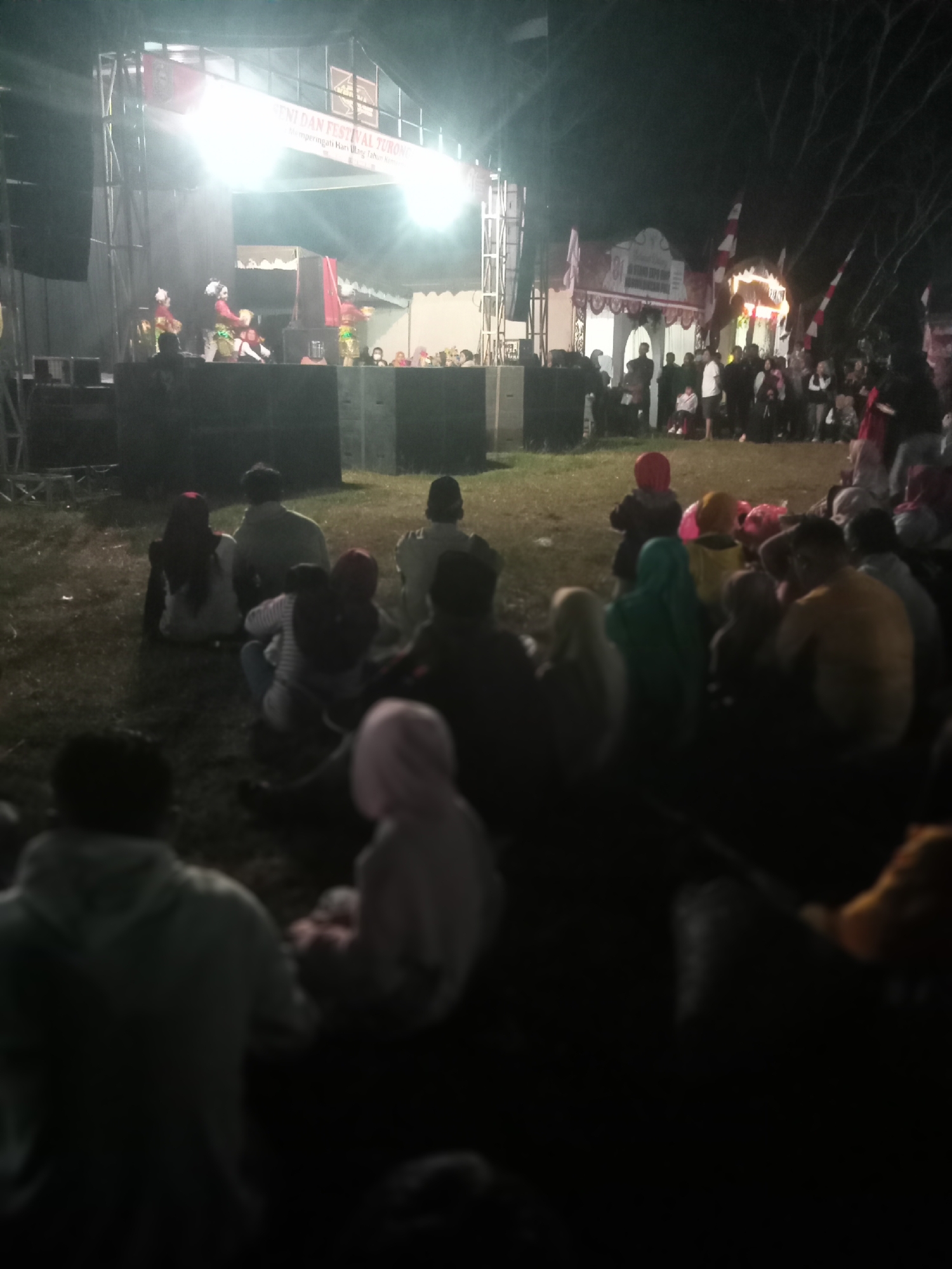 The image depicts an outdoor night event in Pule, Indonesia, possibly a cultural festival or performance. A brightly lit stage is the central focus, featuring performers in traditional attire, likely engaged in a dance or theatrical act. Large speakers are positioned on either side of the stage, and a banner displaying text in Indonesian is visible above the stage, indicating an event titled "SENI DAN FESTIVAL TURONGGO" and commemorating an anniversary.
In front of the stage, a diverse audience is seated on the grassy ground, facing the performers. They are casually dressed, with many illuminated by the stage lights and ambient light from nearby structures. The background shows additional decorated stalls or tents, adorned with red and white flags, suggesting a festive atmosphere. Trees with bare branches are visible in the darker areas of the background, indicating the outdoor setting. The overall scene conveys a lively community gathering and cultural celebration.