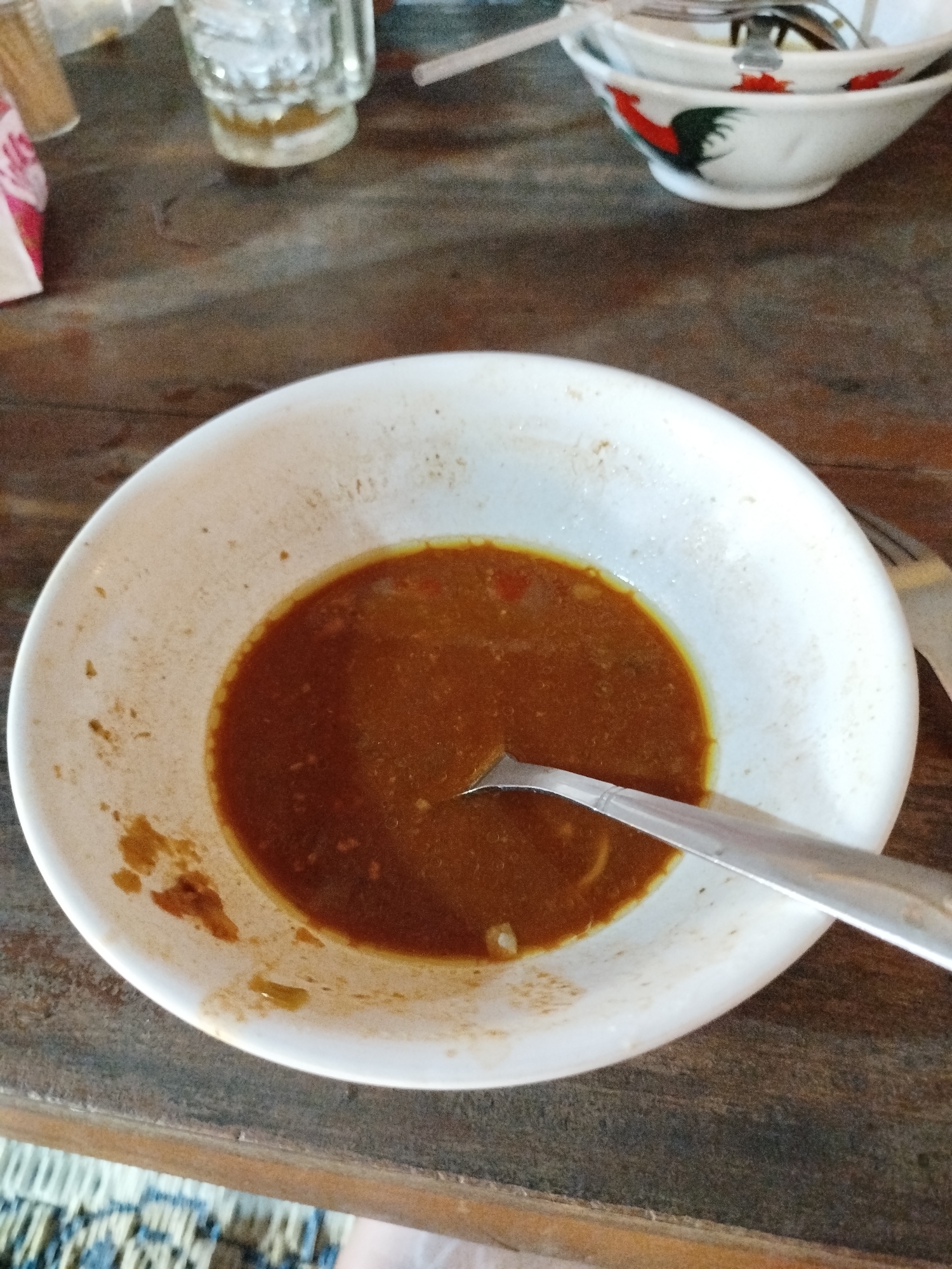 A high-angle, medium shot shows a white bowl with a small amount of brownish-red sauce on a wooden table. A metal spoon rests in the sauce, partially submerged. The bowl is speckled with dried remnants of food. In the background, a stack of white bowls with red and green rooster designs is visible, along with a glass of liquid with ice and a straw. The table surface appears to be made of dark, weathered wood. A small section of a woven mat and a person's leg are visible at the bottom of the frame. The scene is indoors, likely in a casual dining setting in Pojok, Indonesia, given the rooster motif often found on Indonesian ceramics.