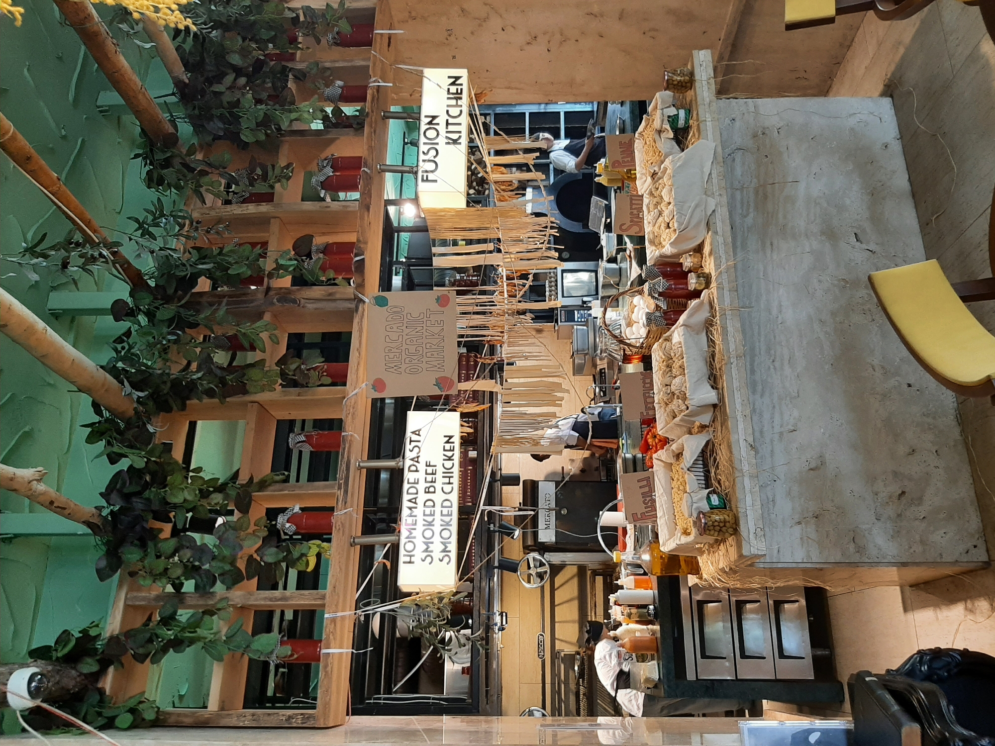 The image shows the interior of a restaurant, likely a pasta shop or a market stall, featuring a rustic and natural aesthetic. Suspended from the ceiling are branches and hanging plants, with red jars placed on wooden shelves. In the foreground, a counter displays various types of pasta in baskets, along with bottles of oil and condiments, and jars of olives. Behind the counter, kitchen equipment, including a stove and ovens, is visible, with chefs in uniform preparing food. Two illuminated signs are prominently displayed: one reads "HOMEMADE PASTA SMOKED BEEF SMOKED CHICKEN," and another, "FUSION KITCHEN." The overall impression is one of fresh, artisanal food preparation in a visually appealing, natural setting. The presence of signs in English and the style of the establishment suggest a tourist-friendly location. While the image itself does not explicitly state the location as Alanya, Turkey, the description of the restaurant and its offerings, combined with the common use of English in tourist areas of Turkey, makes it a plausible interpretation.