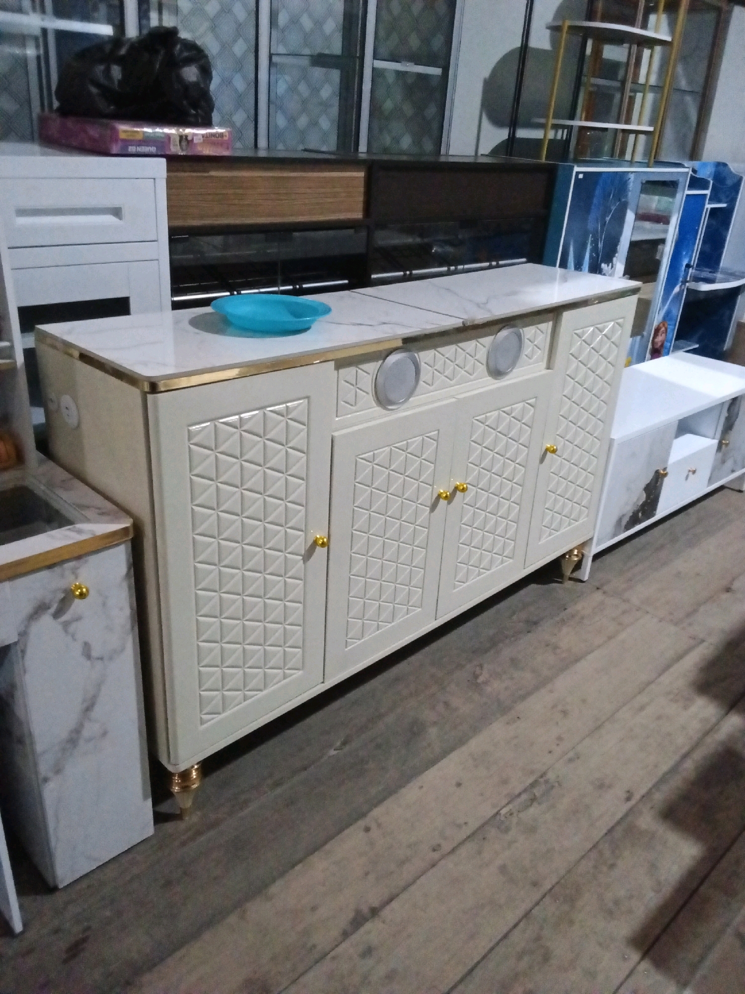 The image depicts a retail display of furniture, likely in a store or showroom located in Gas Alam Badak I, Indonesia. The foreground is dominated by a cream-colored cabinet with intricate geometric patterns on its doors and brass accents. This cabinet appears to be a kitchen island or buffet. To its left, a portion of a white cabinet with a marble-effect countertop and brass trim is visible. In the background, several other furniture pieces are displayed, including shelves and cabinets with blue and white designs, as well as a white TV stand with marble-effect detailing. The flooring consists of aged wooden planks. The scene is well-lit, suggesting an indoor setting. There are no people visible in the image.