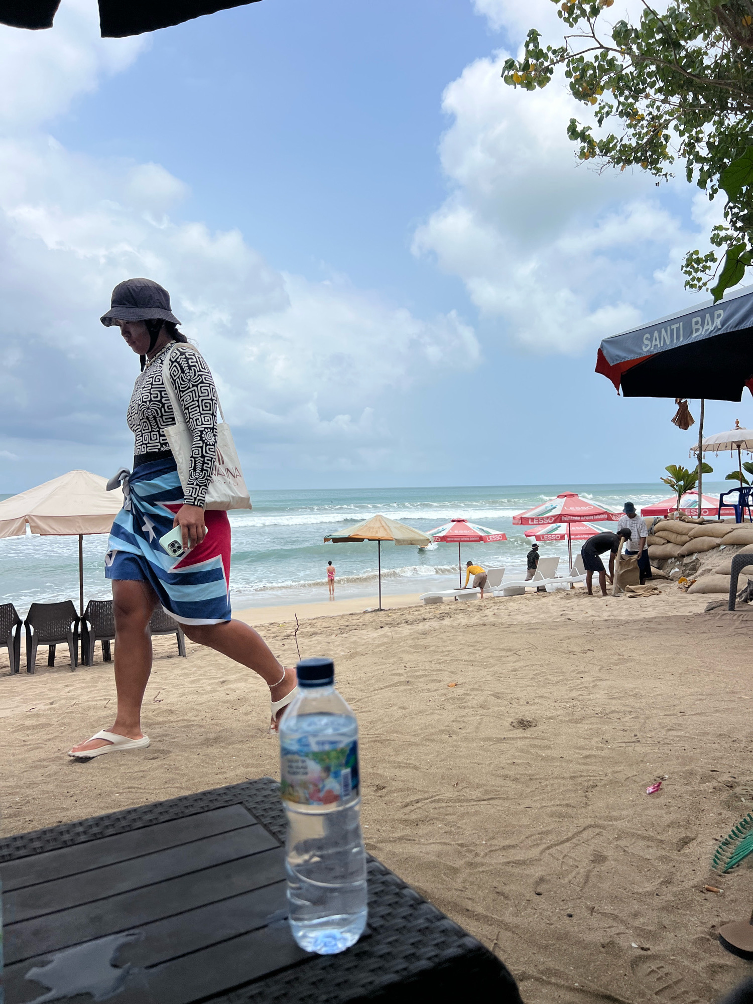 A beach scene in Kuta, Indonesia, shows a woman in a patterned shirt, bucket hat, and a sarong walking on the sand. She is carrying a tote bag and a mobile phone. In the background, several beach umbrellas, lounge chairs, and sandbags are visible. Some individuals are working with sandbags, while others are relaxing or swimming in the ocean. A bottle of water sits on a table in the foreground. The sky is cloudy with patches of blue.