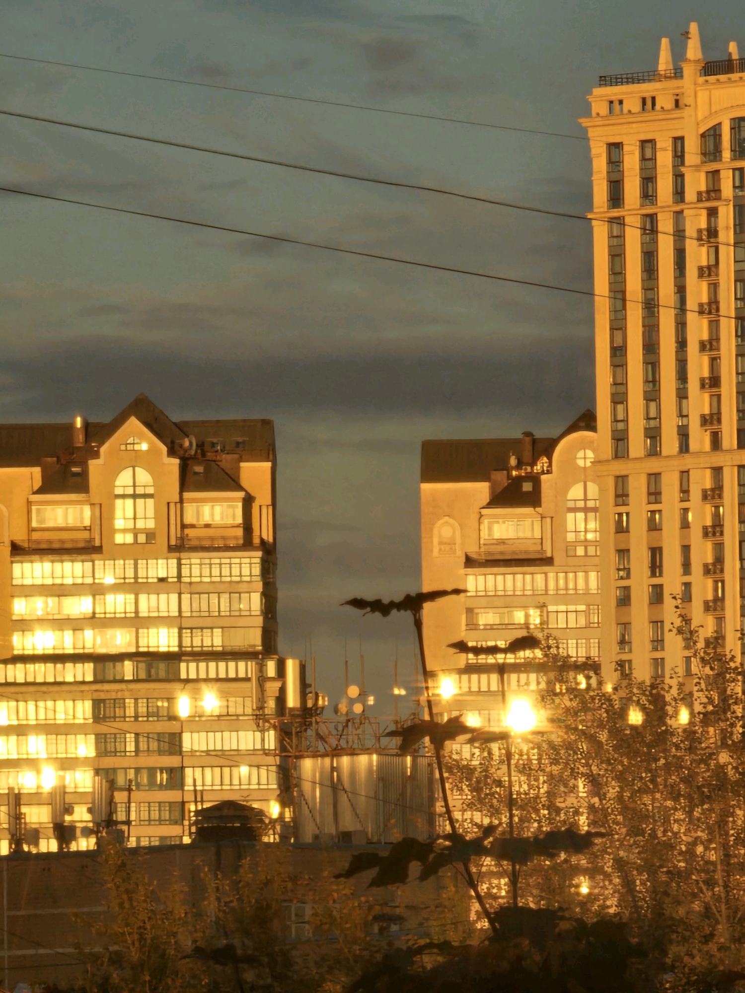The image captures an urban twilight scene in Khimki, Russia, dominated by illuminated modern buildings. In the foreground, silhouetted branches of trees and foliage are visible against the warm glow of artificial lights. Behind them, two multi-story residential buildings are brightly lit from within, their windows casting a golden hue on the facade and reflecting the sky. The building on the right is a taller, more slender structure with a distinctive classical architectural style, featuring vertical window arrangements and decorative elements at the top. The building on the left is shorter and wider, with a more angular roofline and a prominent arched window on its upper level. Power lines stretch across the dusky sky, a common element in urban landscapes. The overall atmosphere is one of quiet evening ambiance, highlighting the contrast between the natural elements of the trees and the man-made structures of the city.